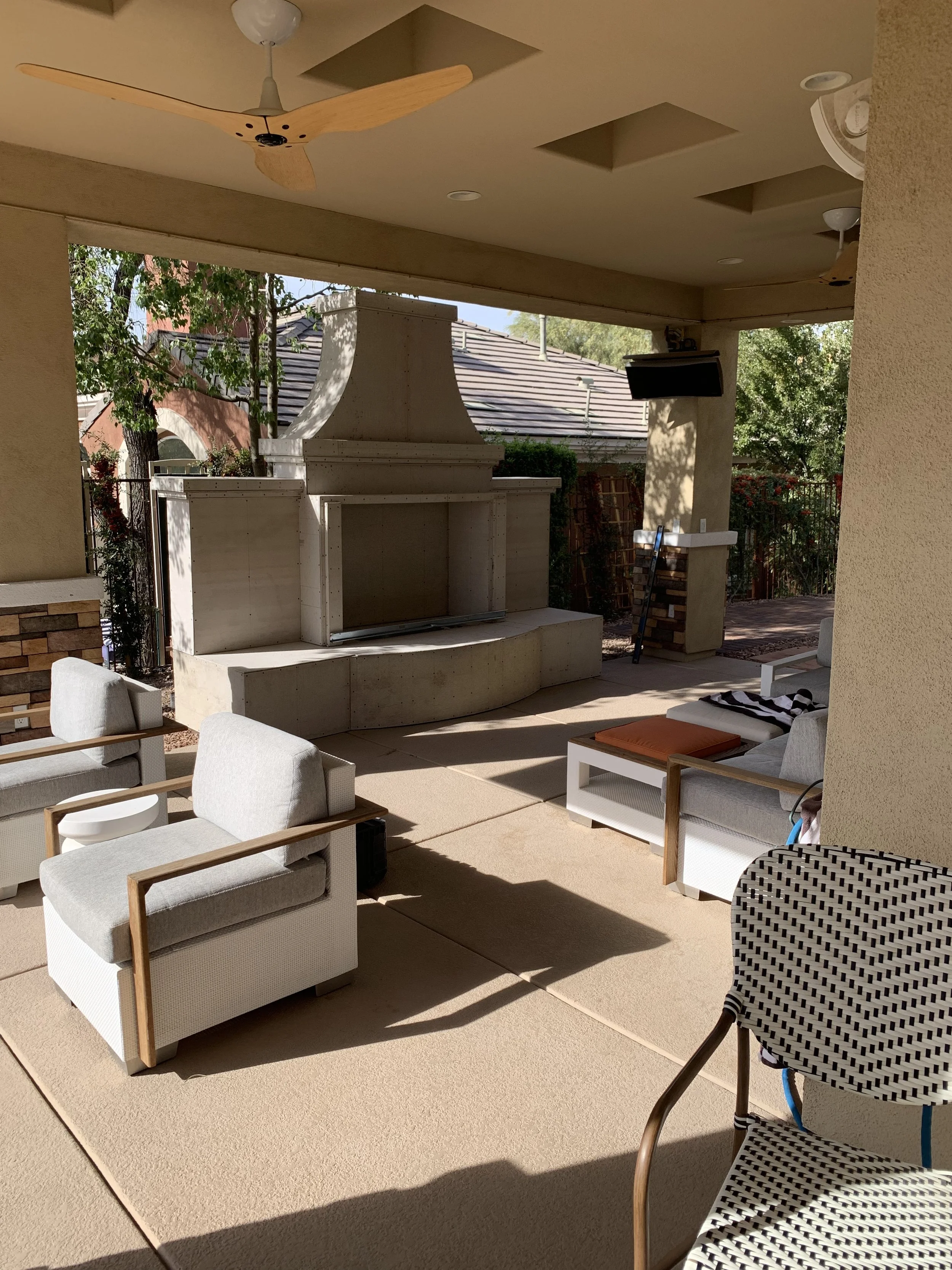 Outdoor patio area with beige furnishings, a fireplace, a ceiling fan, and greenery in the background.