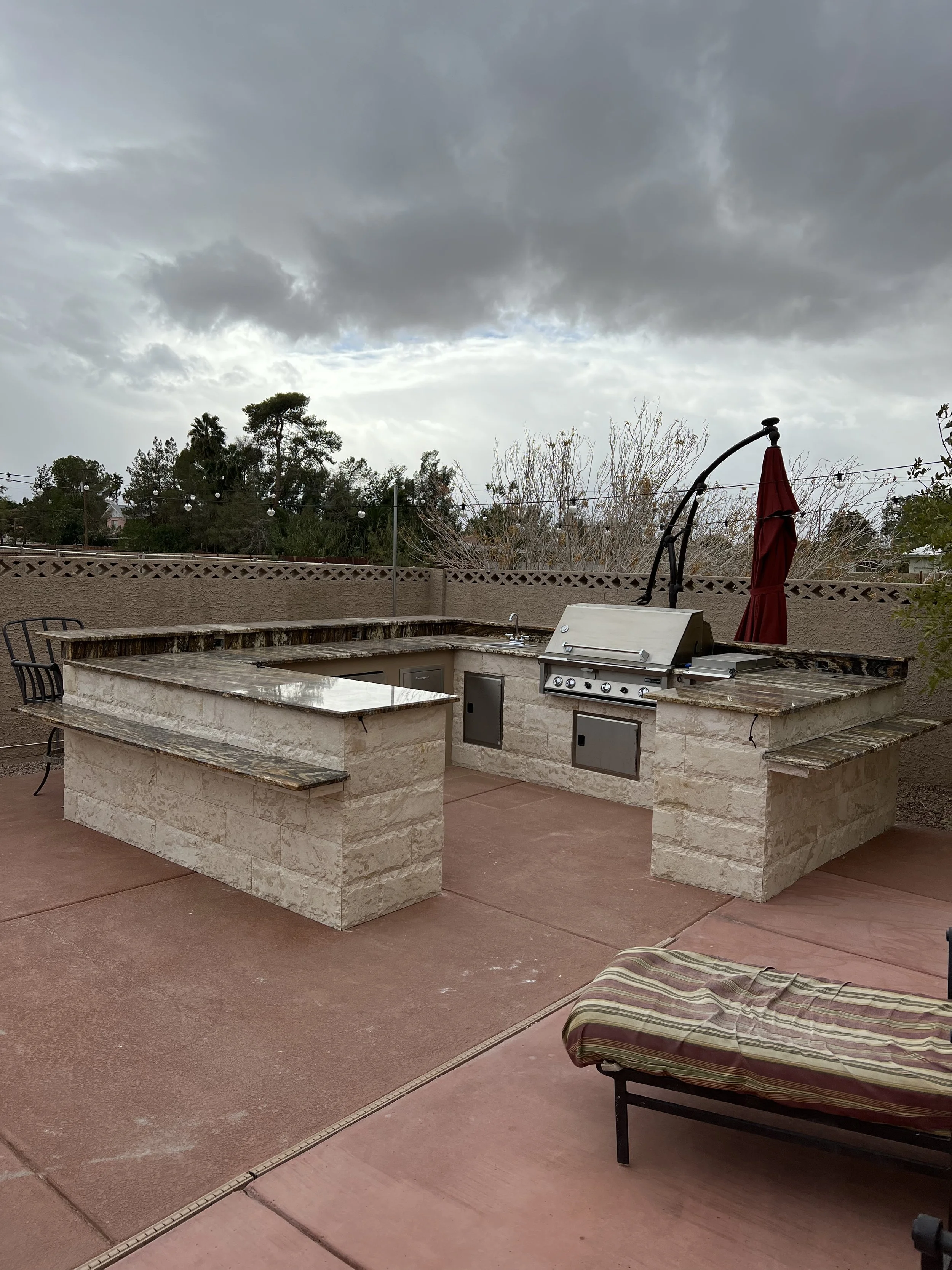 Outdoor kitchen with grill and bar area on a patio, stone construction, overcast sky.
