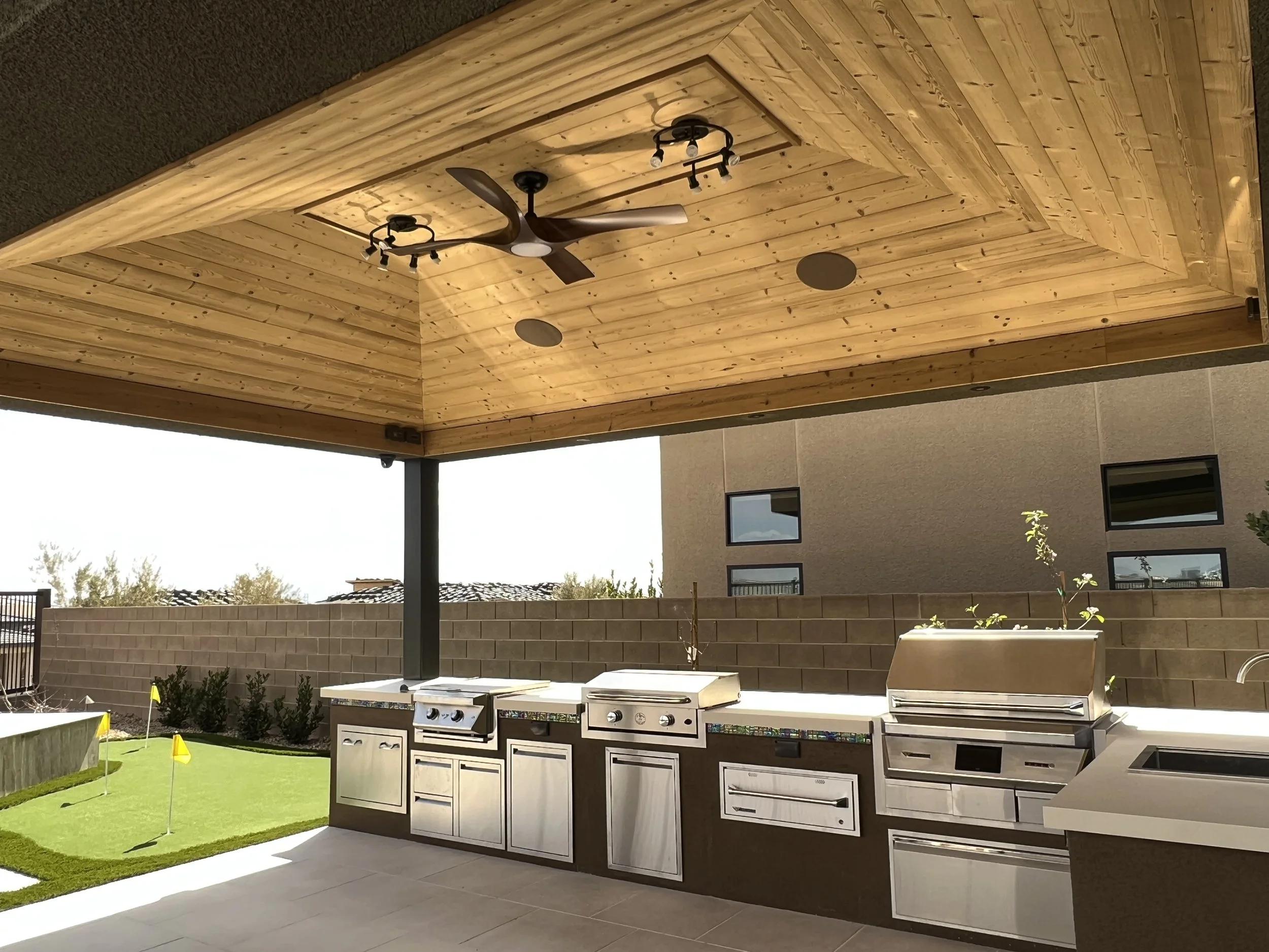 Covered outdoor kitchen with stainless steel appliances, wood paneled ceiling with fan, small putting green with yellow flags, and surrounding brick wall.