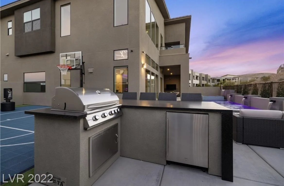 Modern outdoor kitchen with grill and refrigerator on patio of luxury home with basketball hoop and pool at sunset.