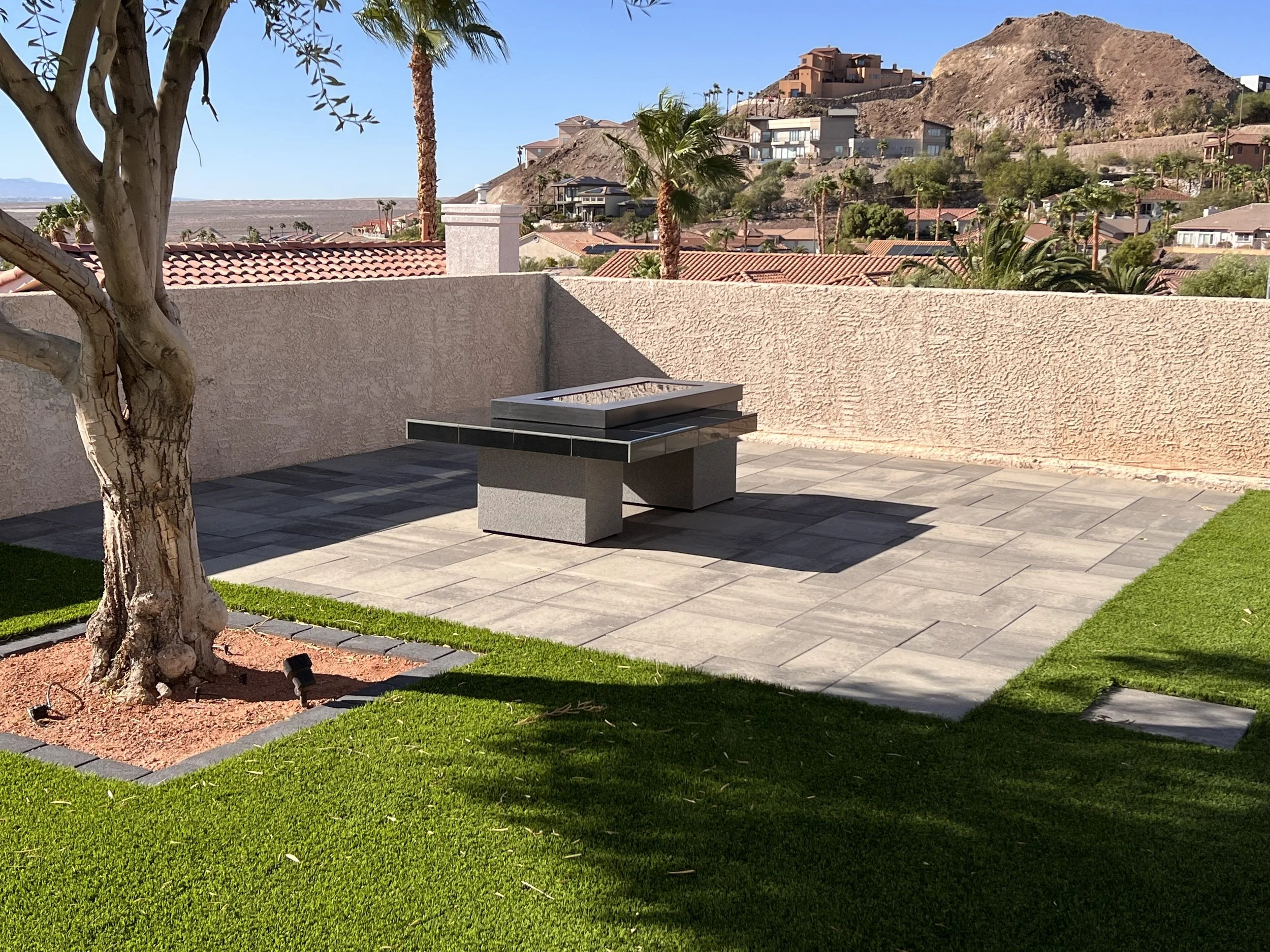 Outdoor patio area with a square fire pit on a tiled surface, surrounded by artificial grass, a tree, and desert landscaping in the background.