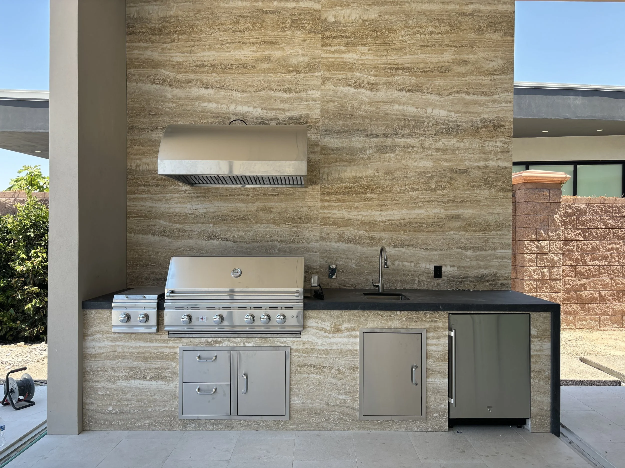 Outdoor kitchen with built-in grill and sink, stainless steel appliances, and stone wall.