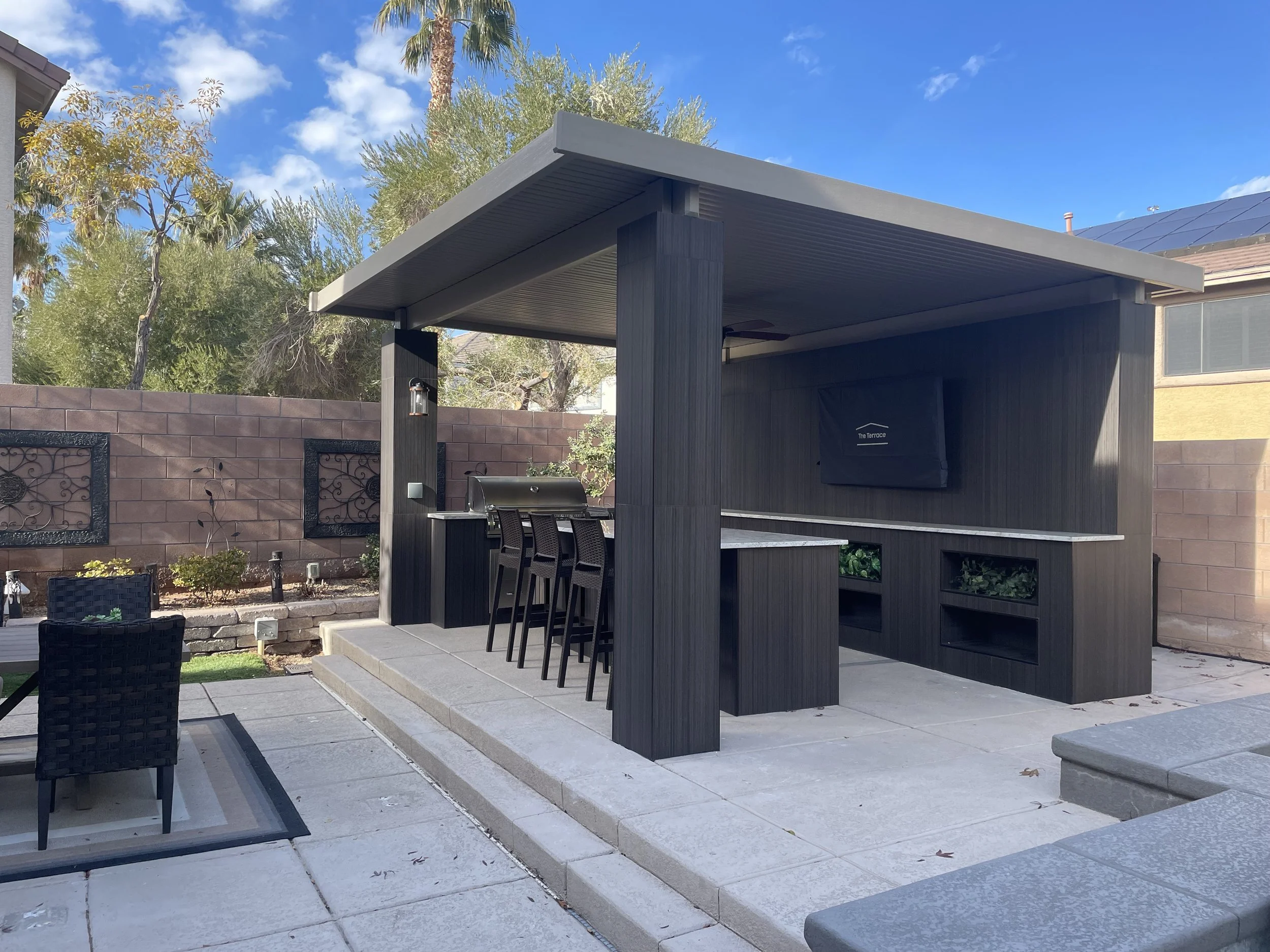 Modern outdoor kitchen with a covered seating area, grill, bar stools, and surrounding garden in a backyard.