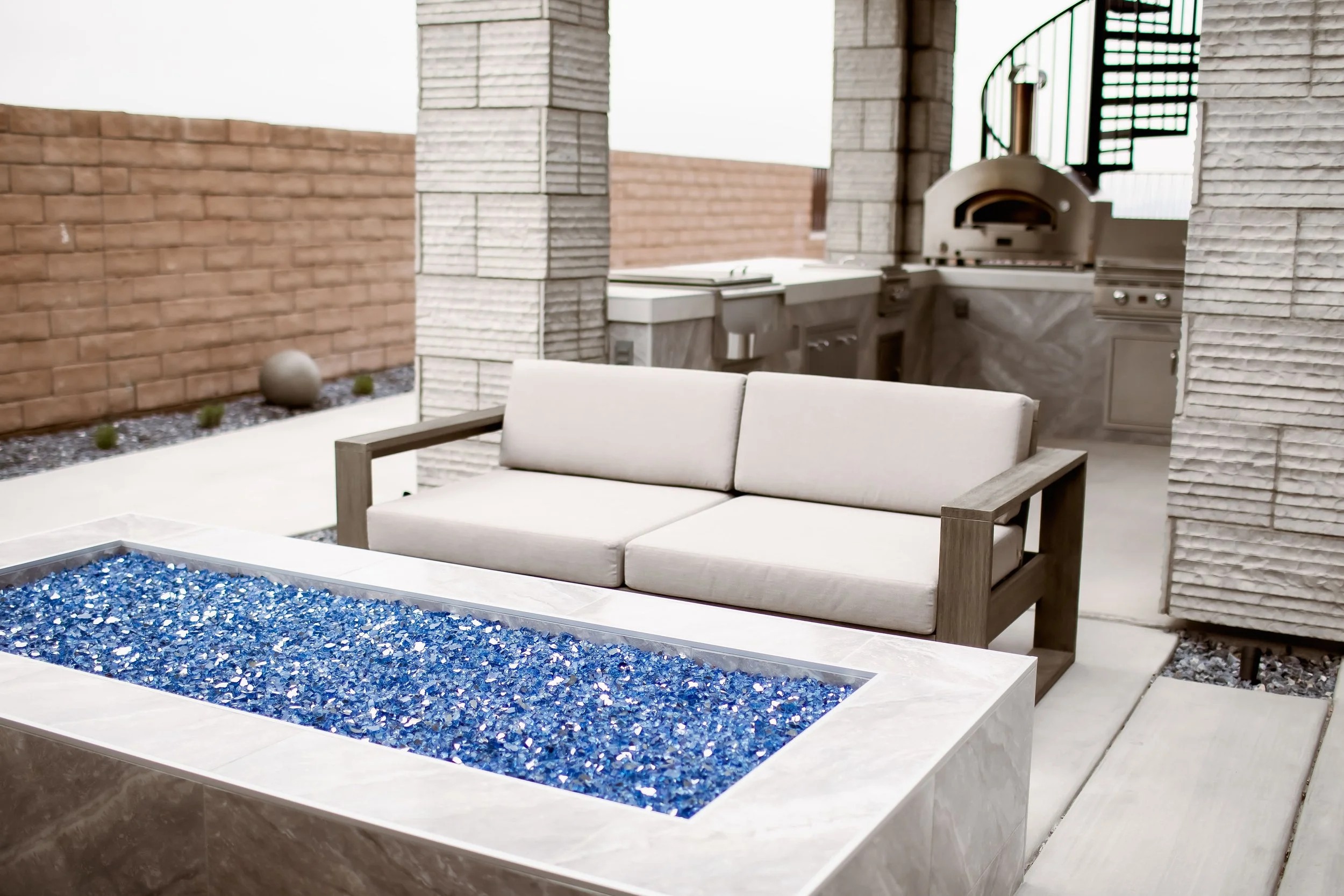 Outdoor patio with a modern sectional sofa, a fire pit with blue glass stones, brick pillars, and an outdoor kitchen with a pizza oven and grill.
