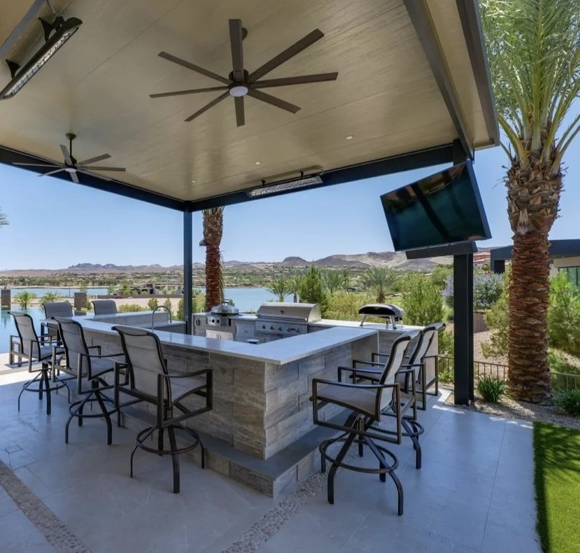 Outdoor kitchen with bar seating, large grill, and ceiling fans under a covered patio, overlooking a scenic landscape with palm trees and distant hills.
