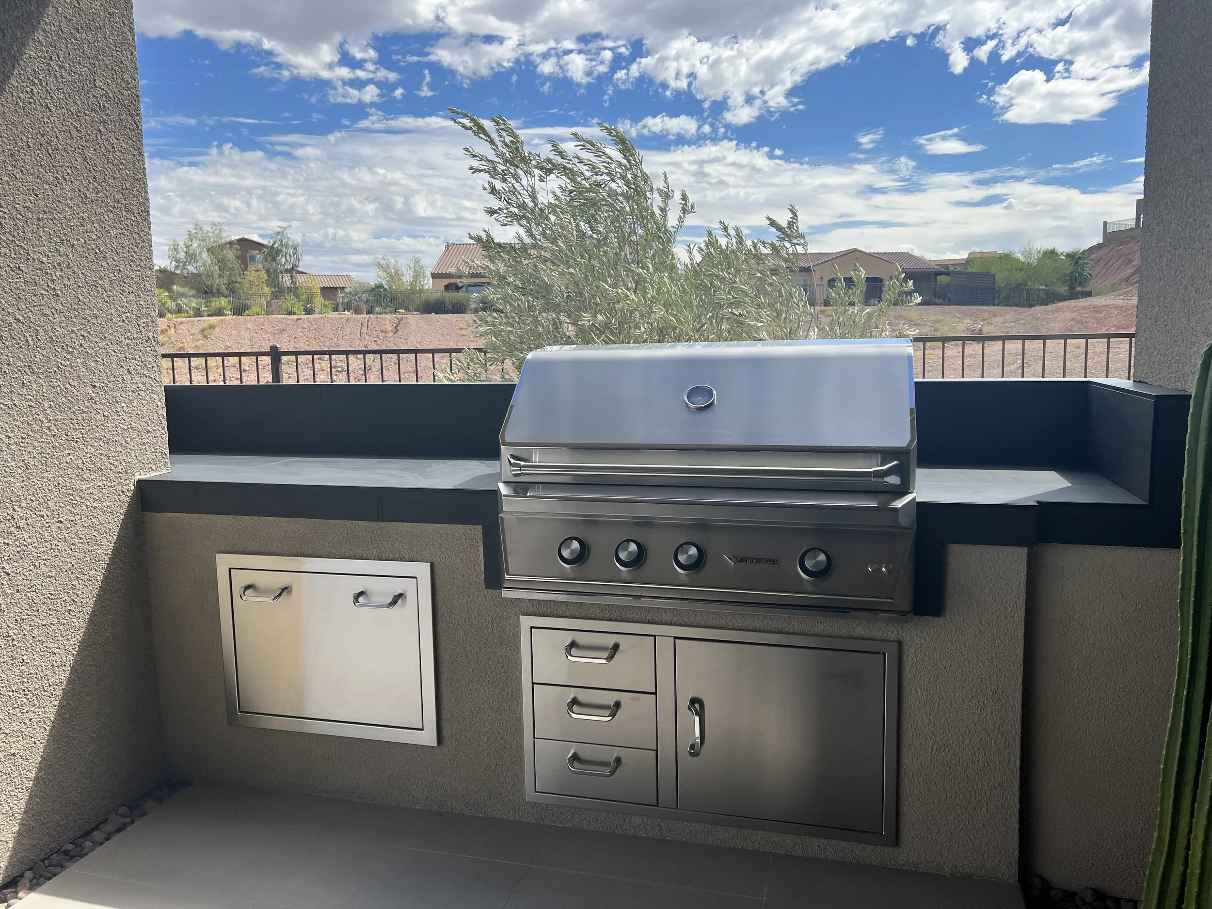 Outdoor built-in stainless steel barbecue grill with storage cabinets, set against a house wall on a patio with a view of a residential neighborhood and cloudy sky.