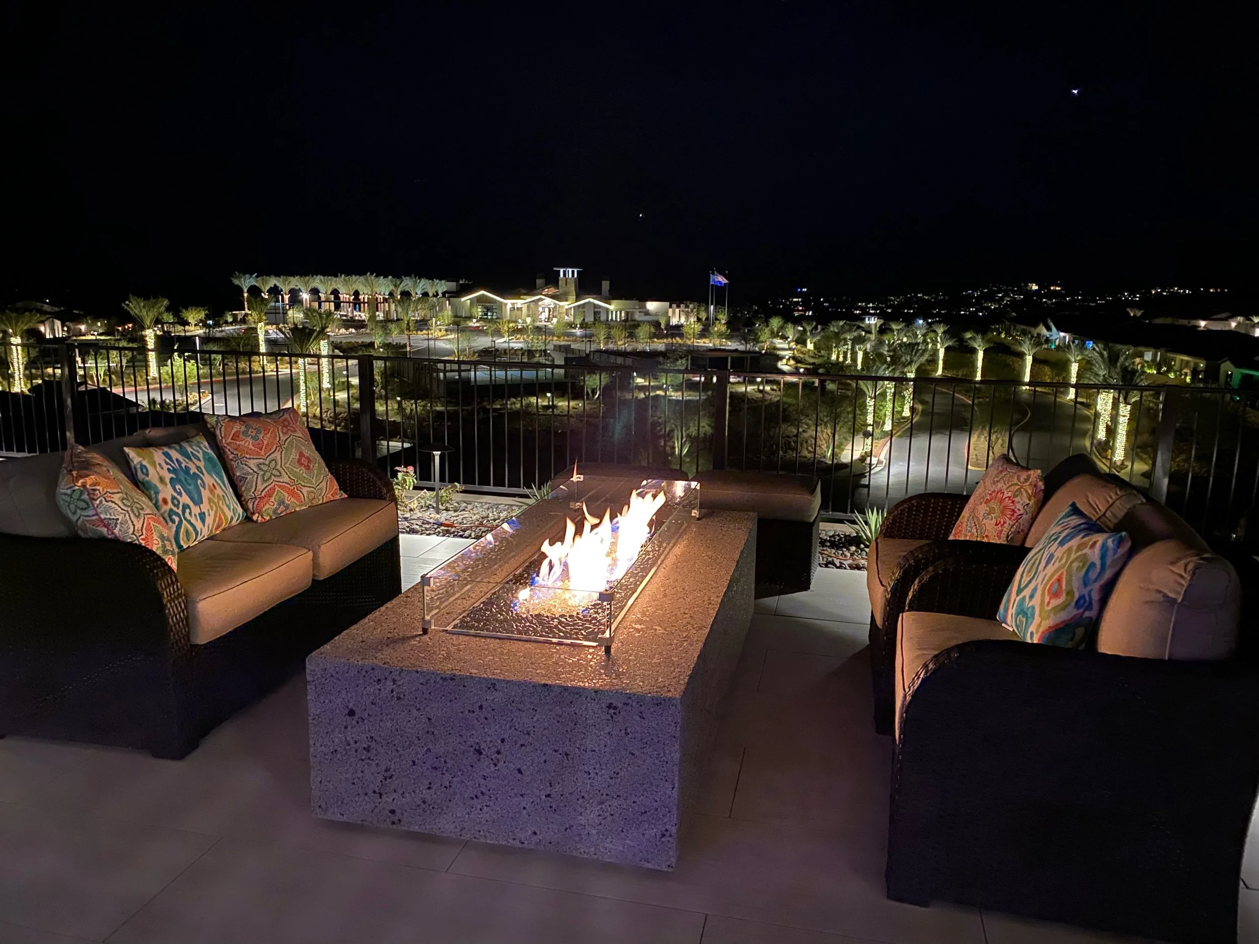 Nighttime patio with two wicker sofas, colorful cushions, and a rectangular fire pit. Background features illuminated garden landscape and distant lights.