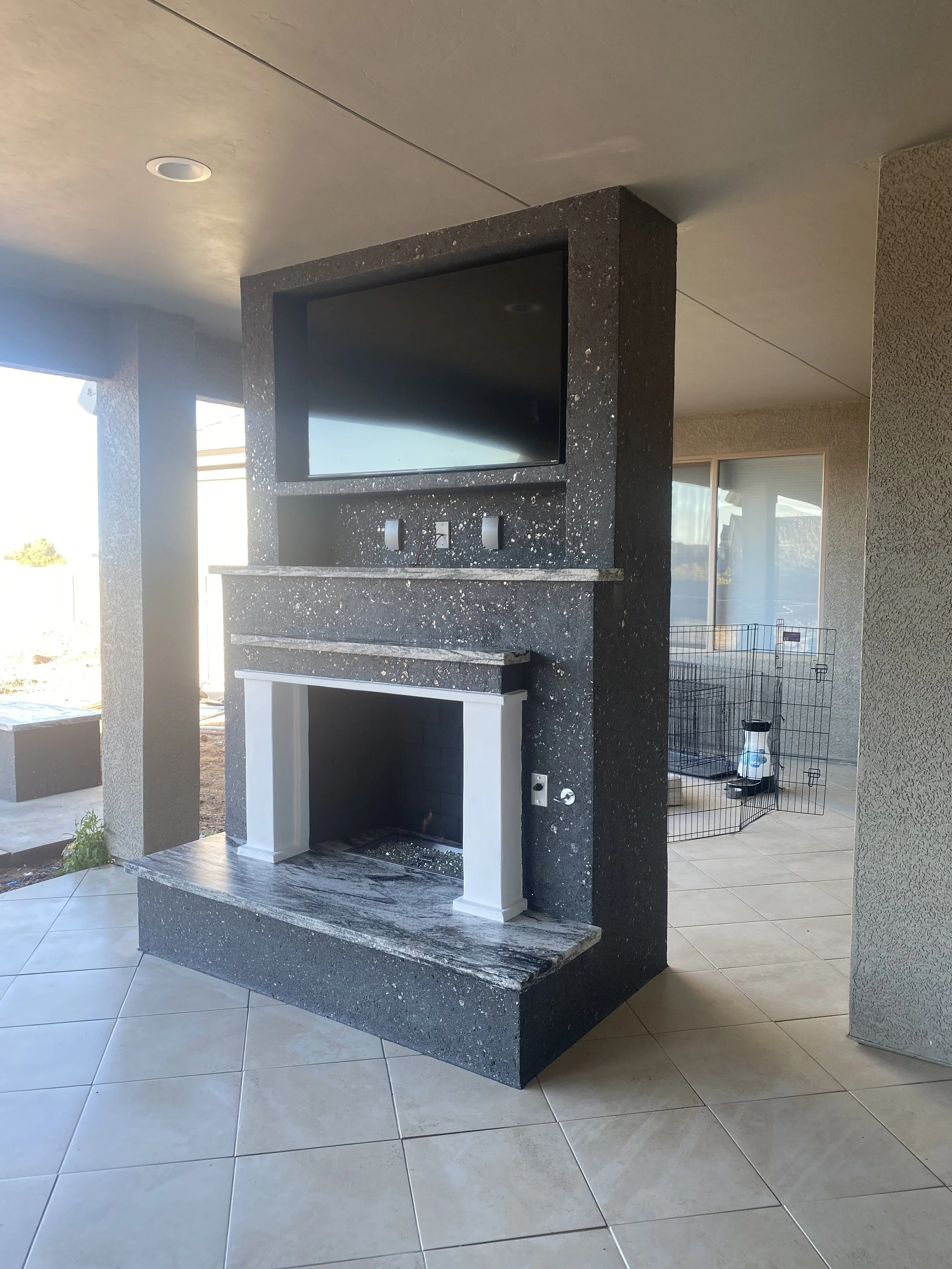 Outdoor patio with a stone fireplace, mounted TV above, tiled floor, and a dog playpen in the background.