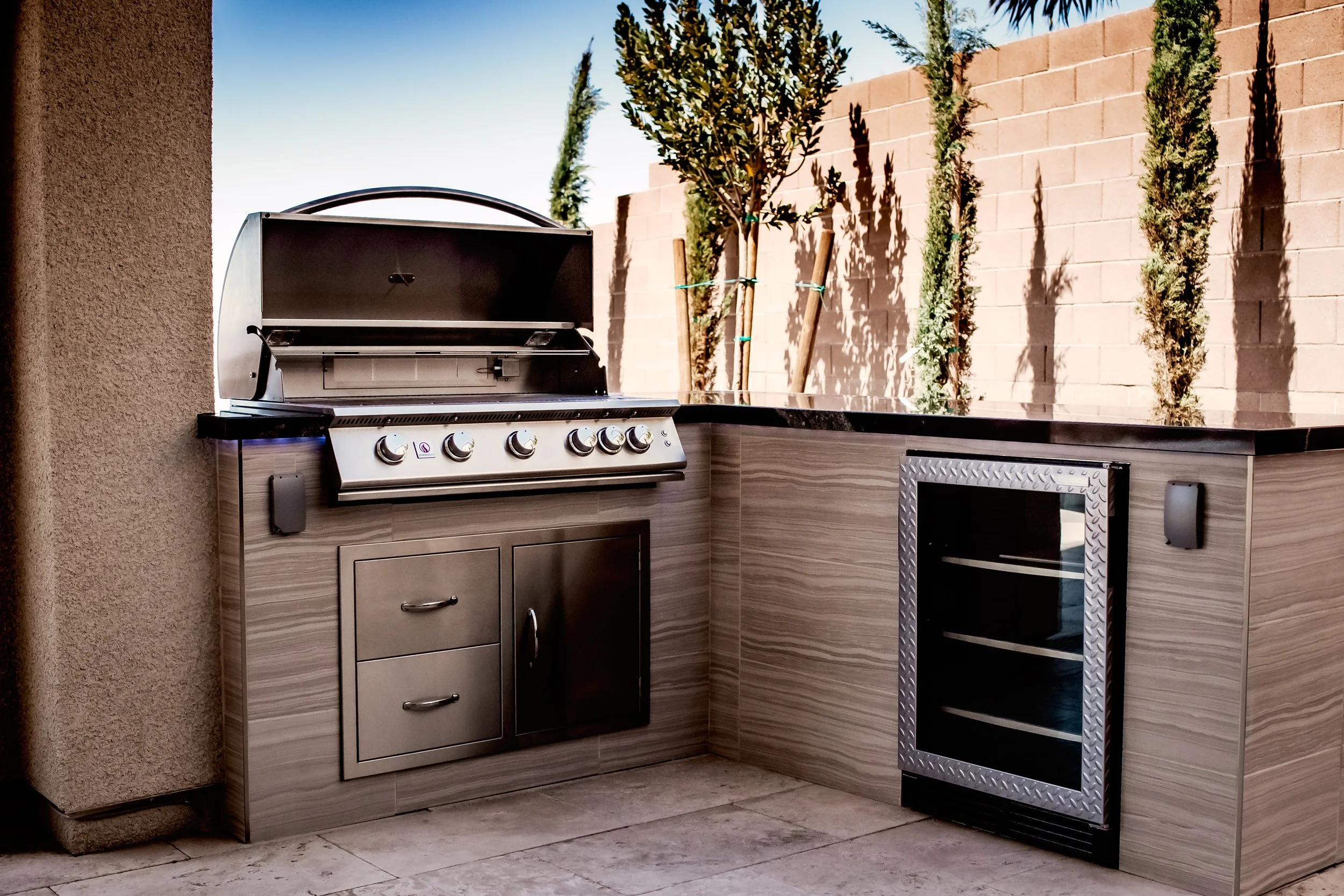 Outdoor kitchen with stainless steel gas grill and mini refrigerator set in a stone countertop, surrounded by a brick wall and small trees.