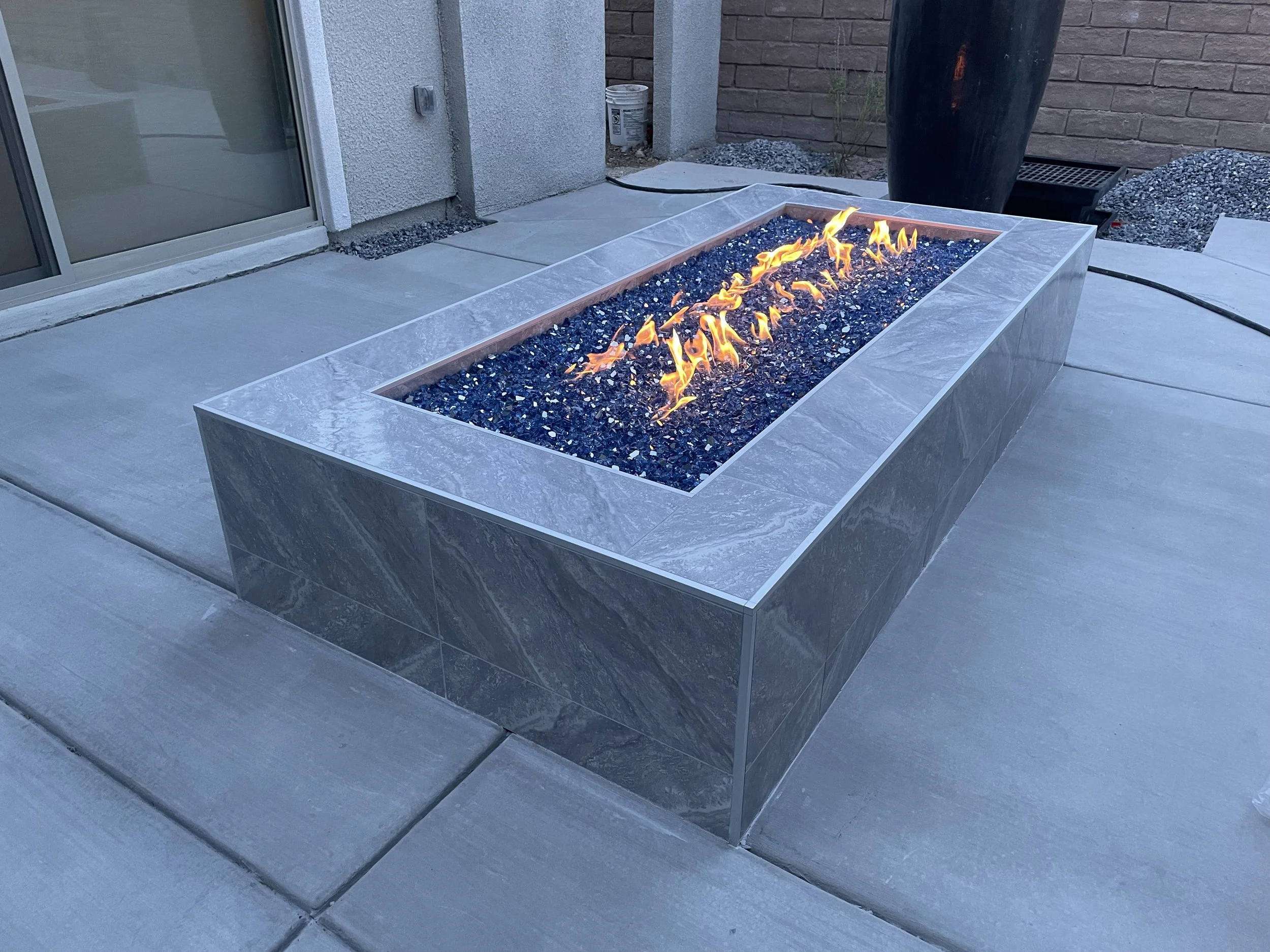 Modern rectangular fire pit with blue glass rocks, lit with flames, on a concrete patio.