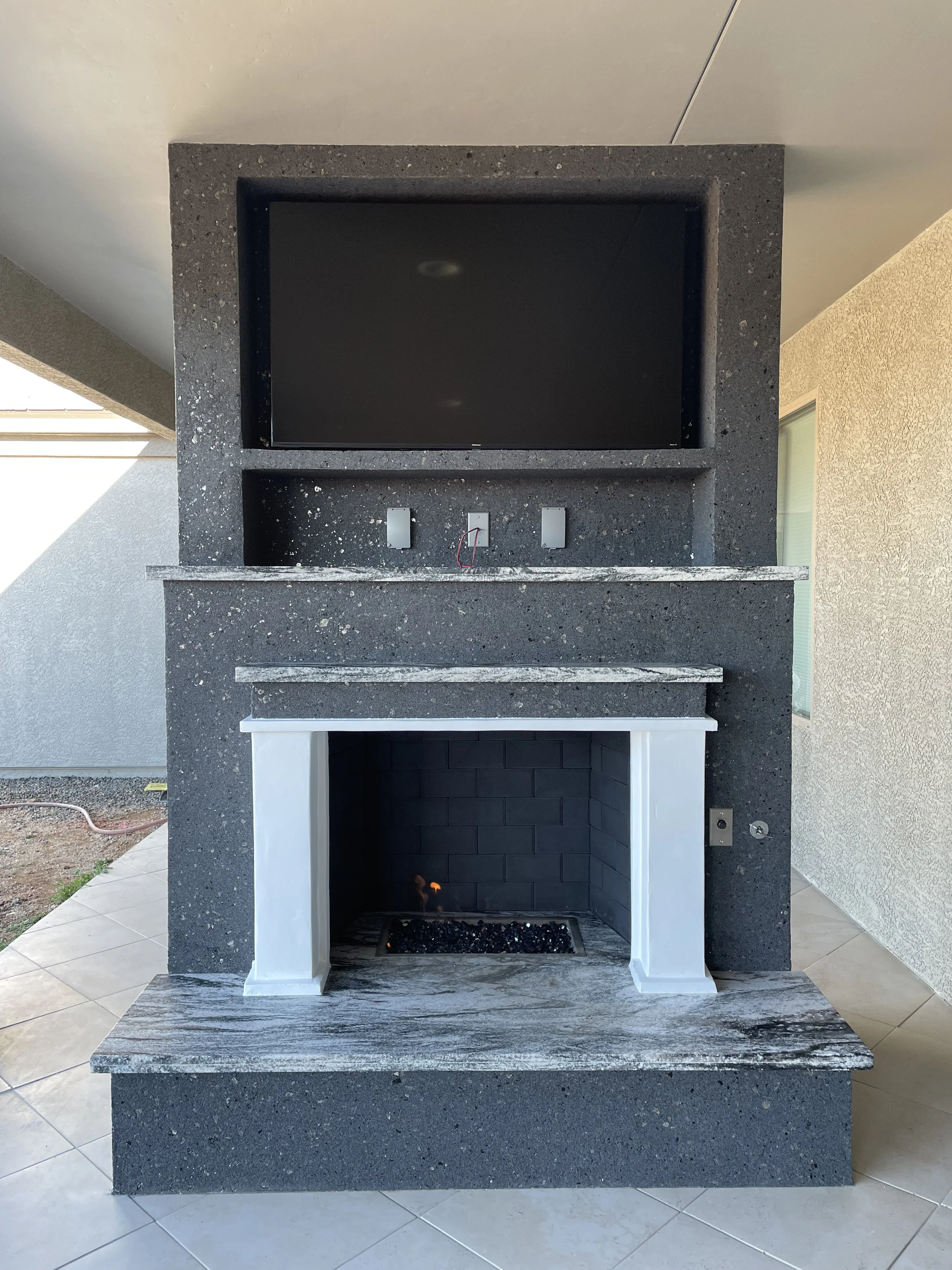 Outdoor entertainment area with a large TV mounted above a modern fireplace, featuring a dark stone surround and a small fire burning in a recessed hearth, tiled patio flooring.