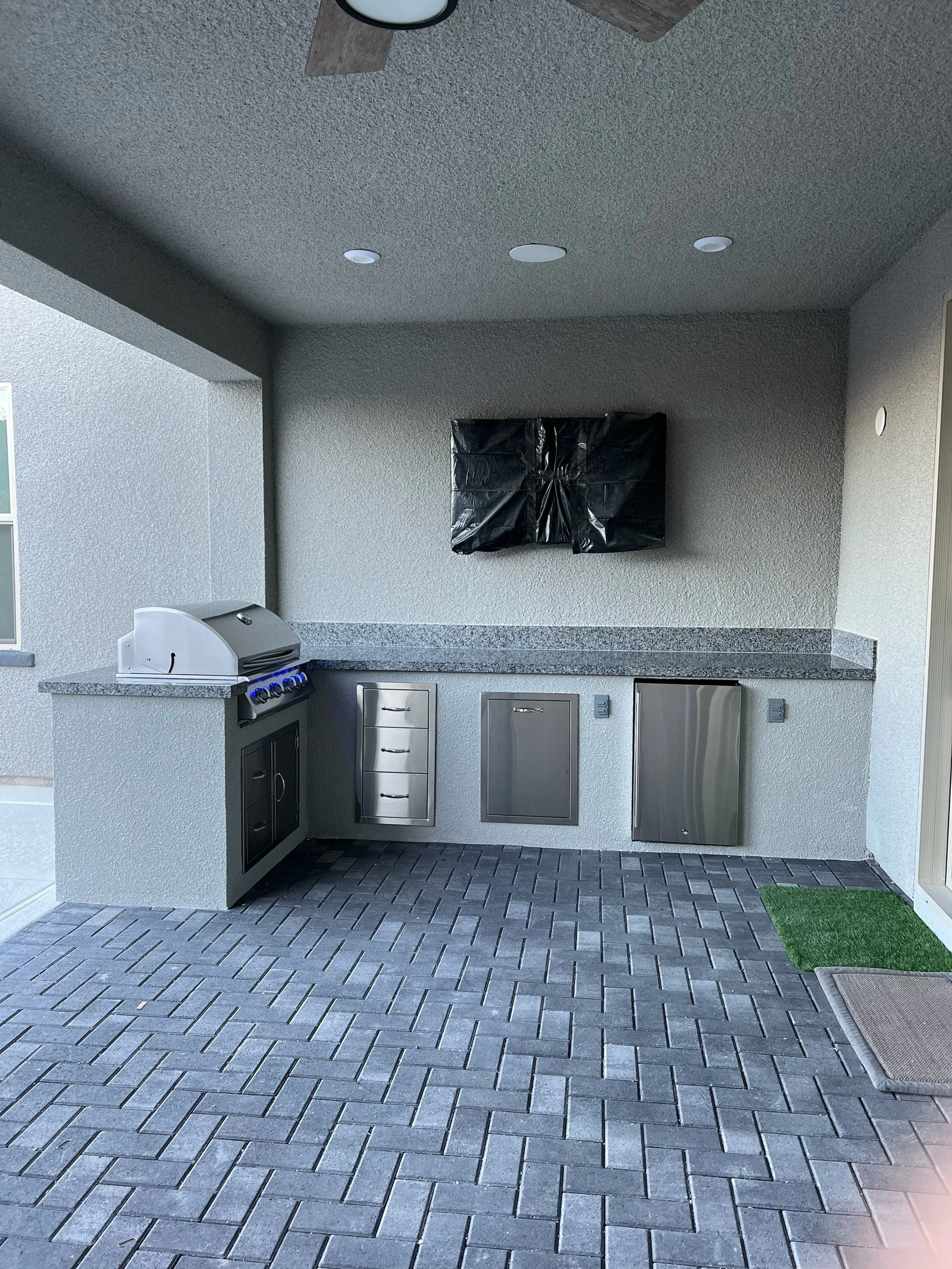 Outdoor kitchen with a built-in grill, countertops, stainless steel drawers, and storage, covered patio ceiling with lights and ceiling fan, wall-mounted covered item, tiled floor, and a small patch of artificial grass.