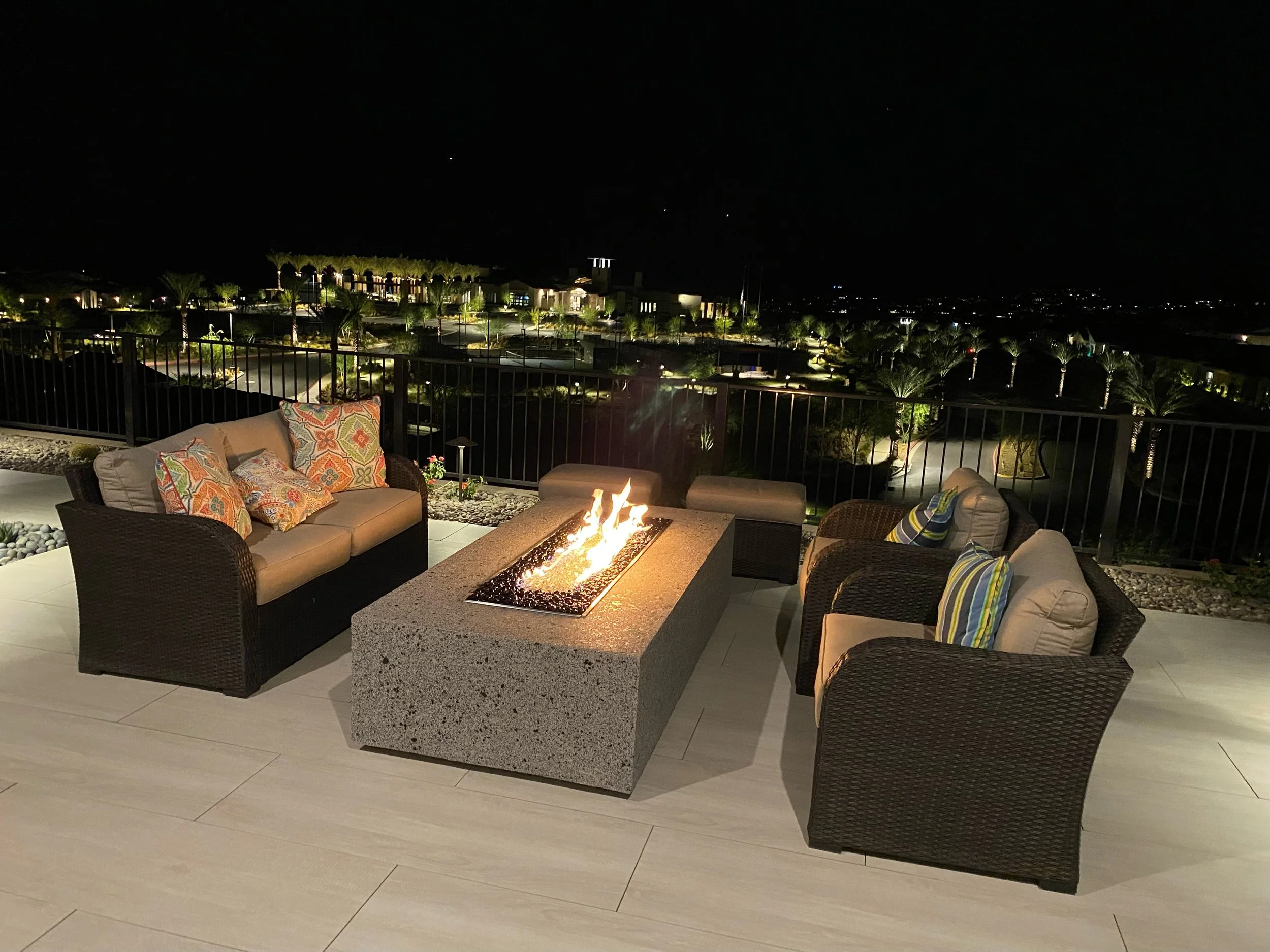 Outdoor patio with wicker furniture set around a rectangular fire pit table, overlooking a landscaped area illuminated at night.