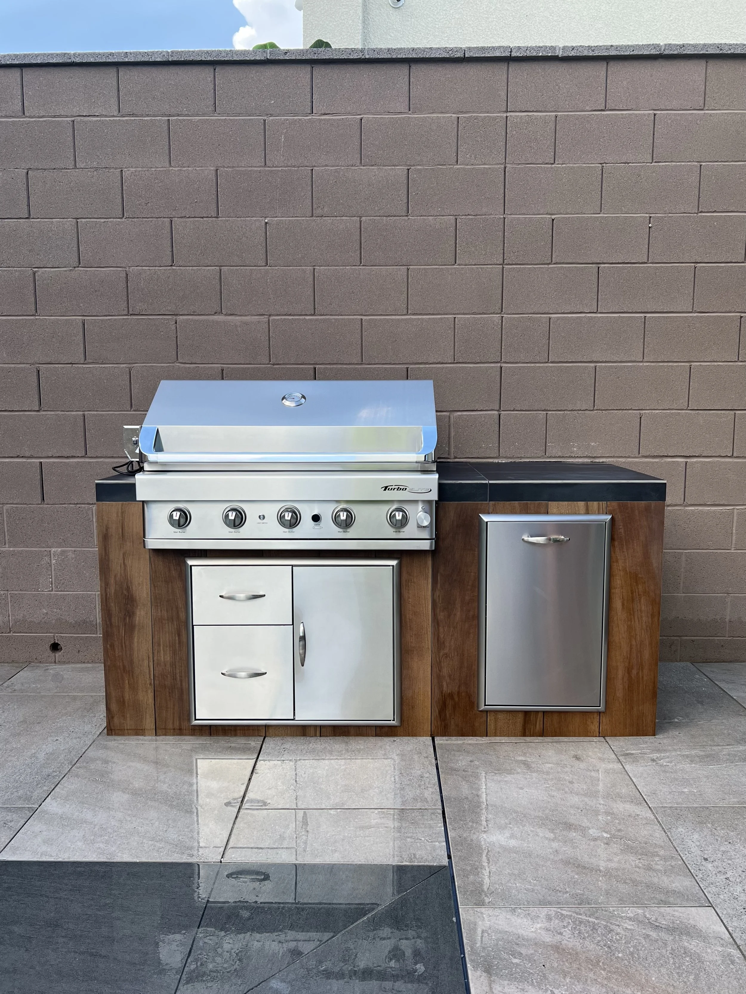 Outdoor stainless steel barbecue grill with wooden base, featuring multiple knobs, drawers, and a side compartment, set against a brick wall on a tiled patio.