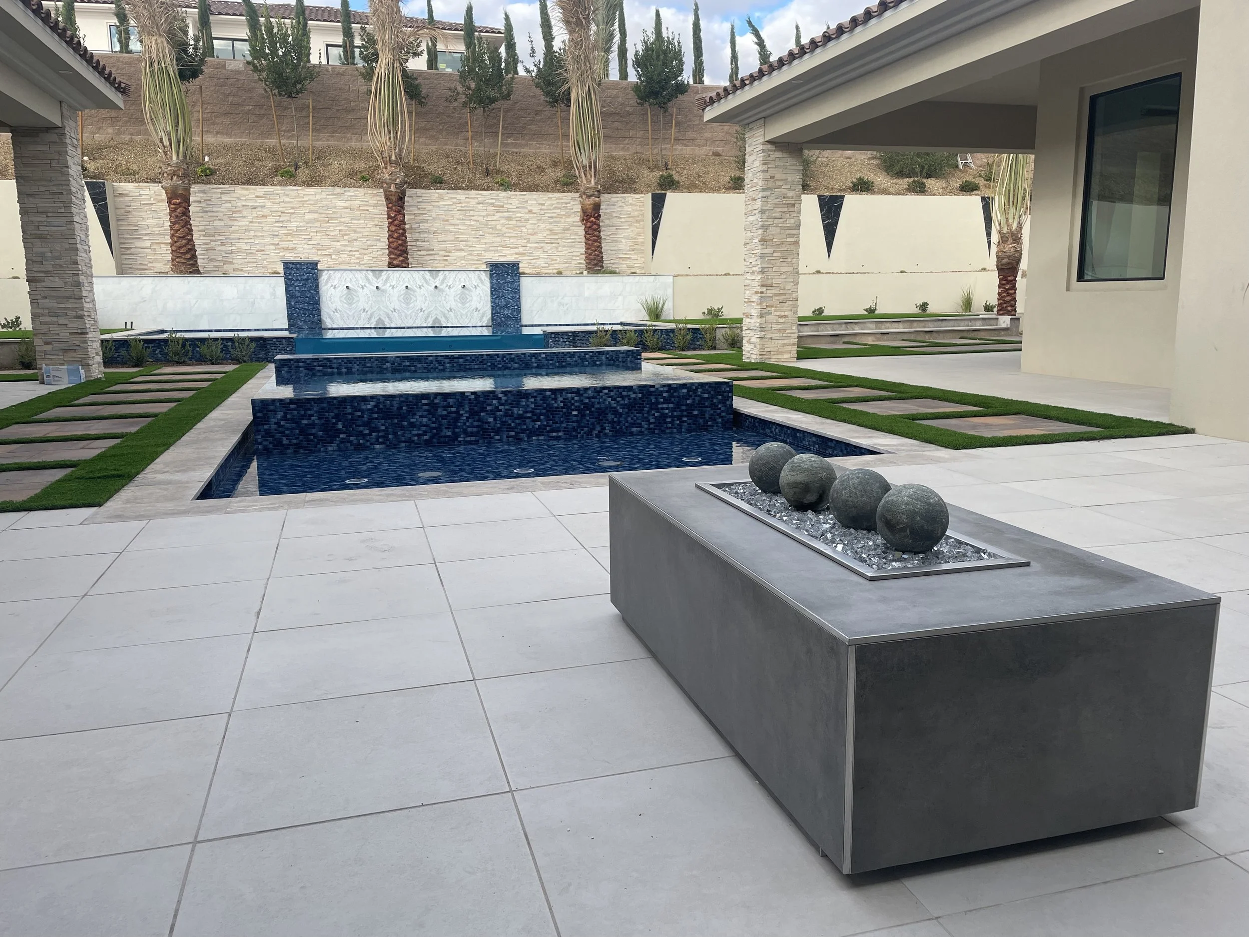 Outdoor patio with modern design, featuring a rectangular water fountain with blue mosaic tiles and decorative stone spheres on a concrete table. There are landscaped areas with stepping stones and grass, as well as a tall stone wall with palm trees.