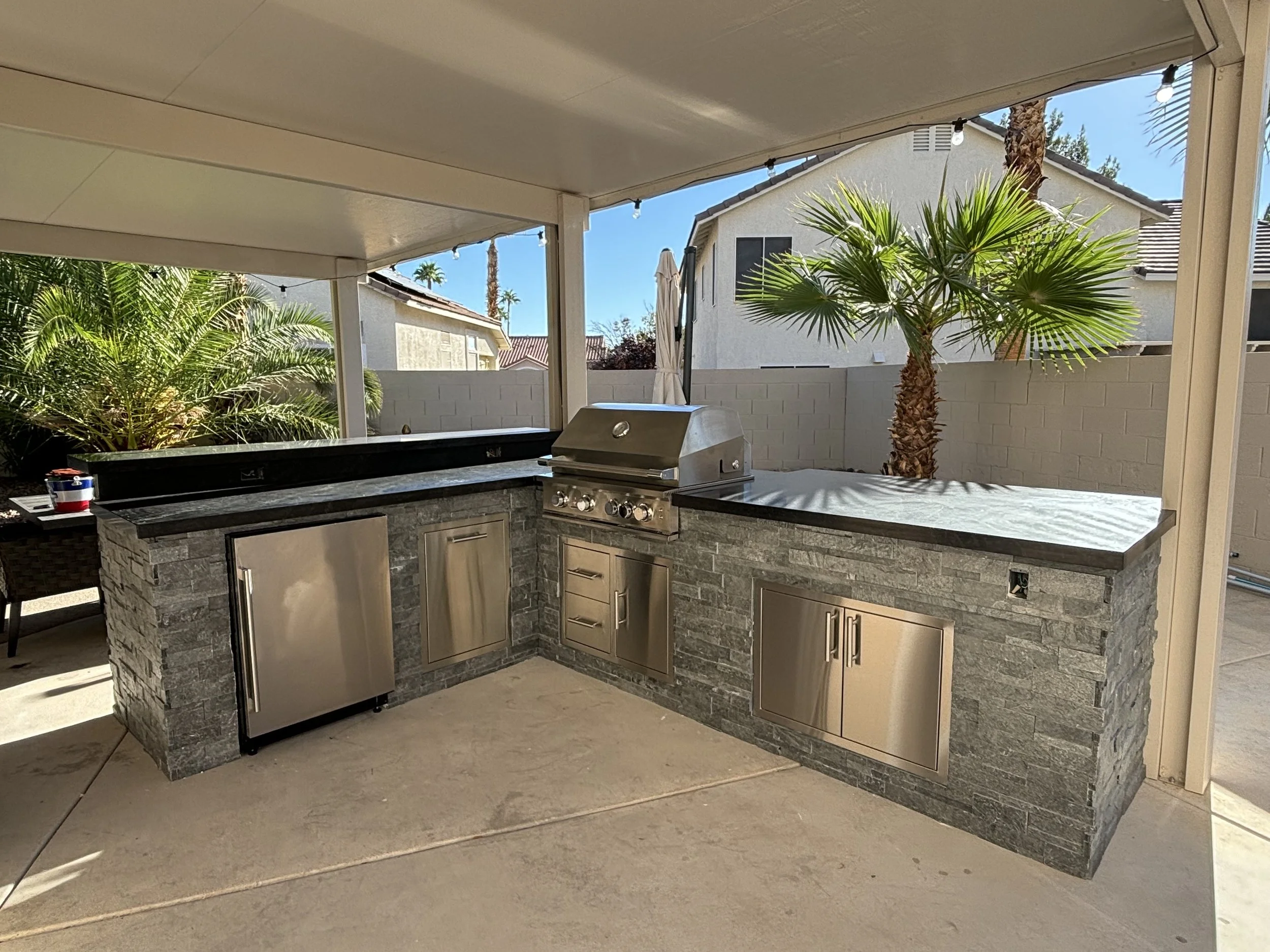 Outdoor kitchen with stainless steel appliances, including a grill and refrigerator, under a covered patio with palm trees visible in the background.