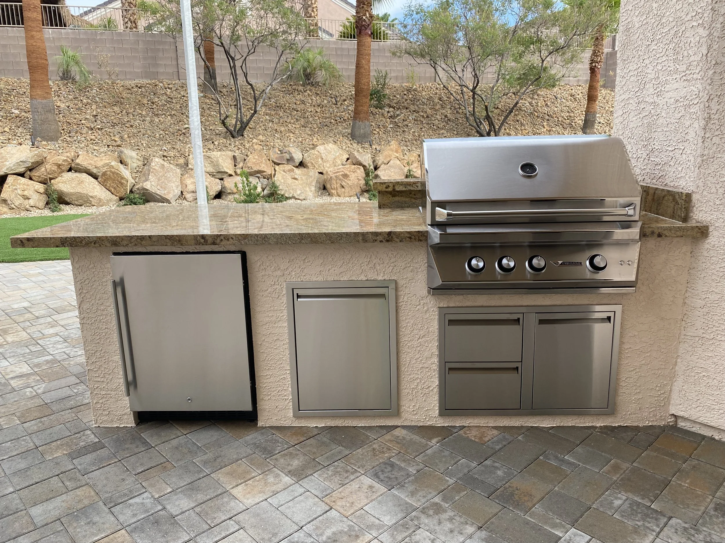 Outdoor kitchen with stainless steel grill, refrigerator, and storage drawers on a stone patio.