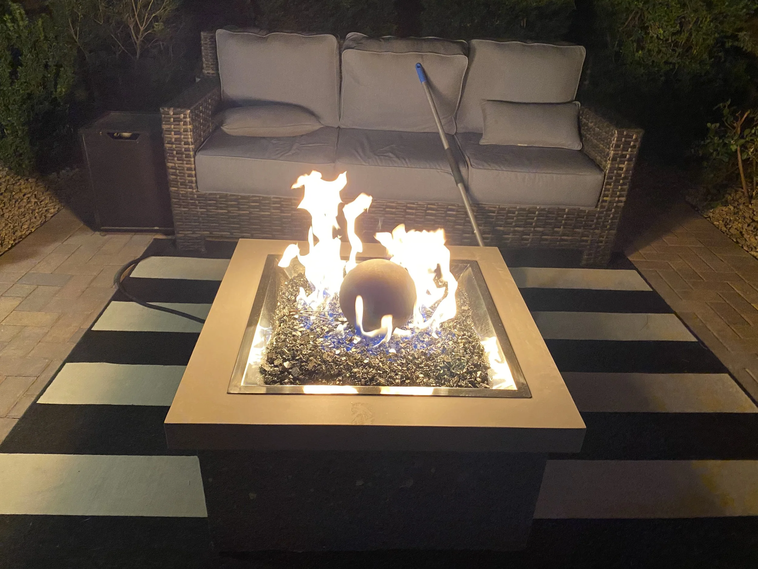 Patio with wicker couch, fire pit table with flames, striped rug, and a garden hoe.