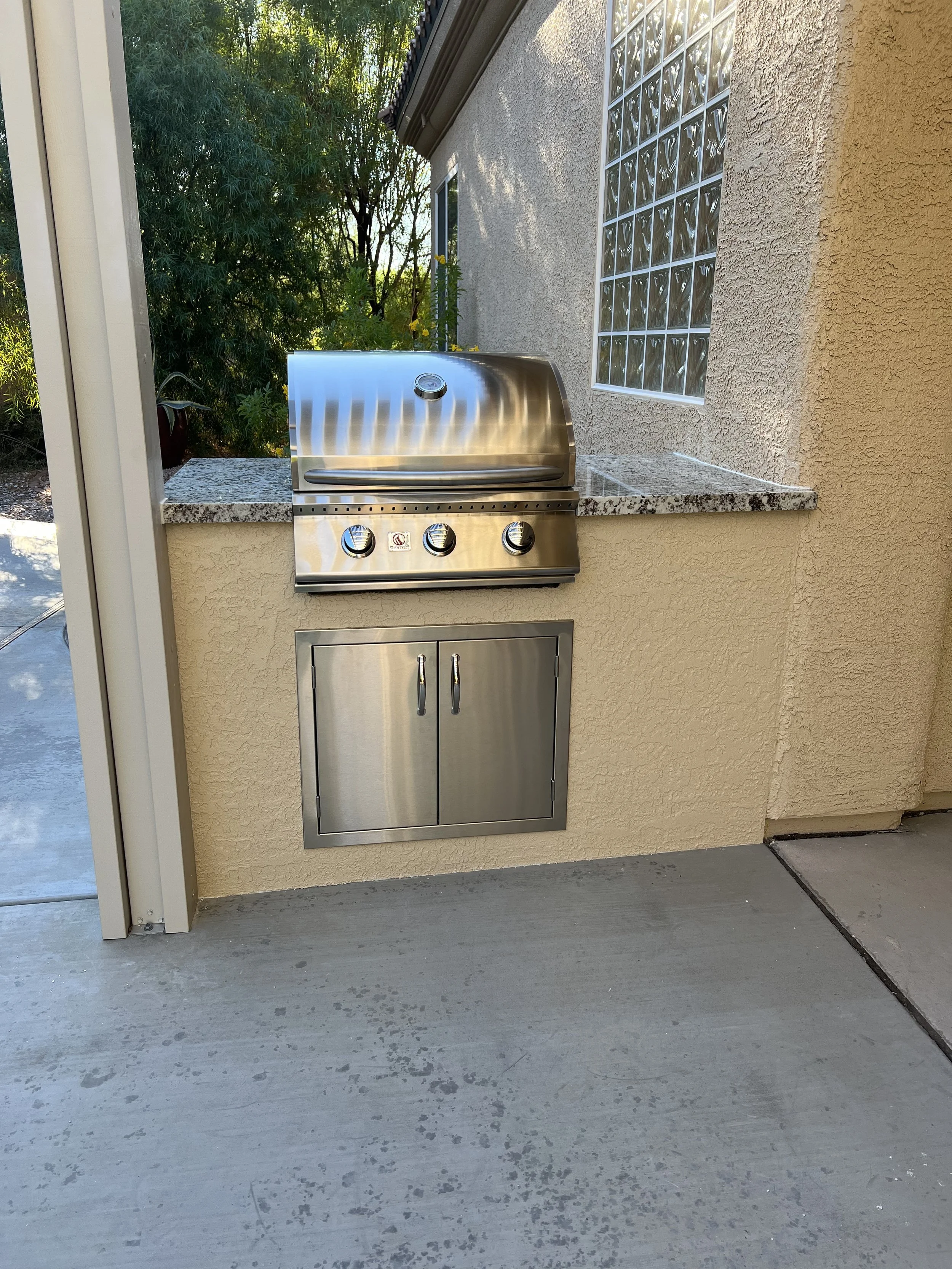 Outdoor stainless steel built-in grill with closed lid and storage cabinet underneath, set against a stucco wall with a window and surrounded by greenery.