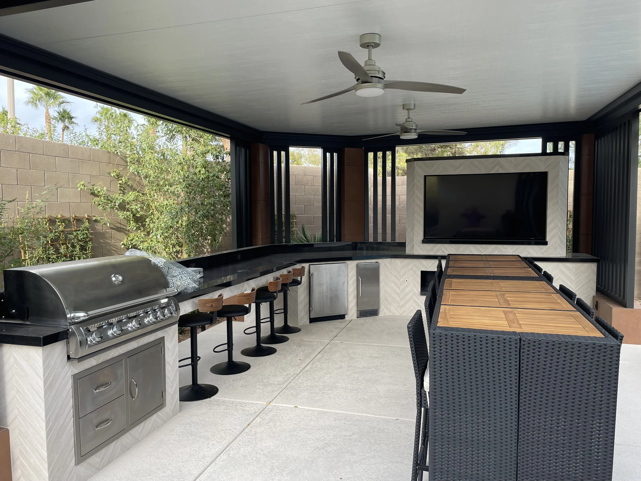 Outdoor patio with stainless steel barbecue grill, bar stools, rattan dining set, large TV, overhead fans, and plants.