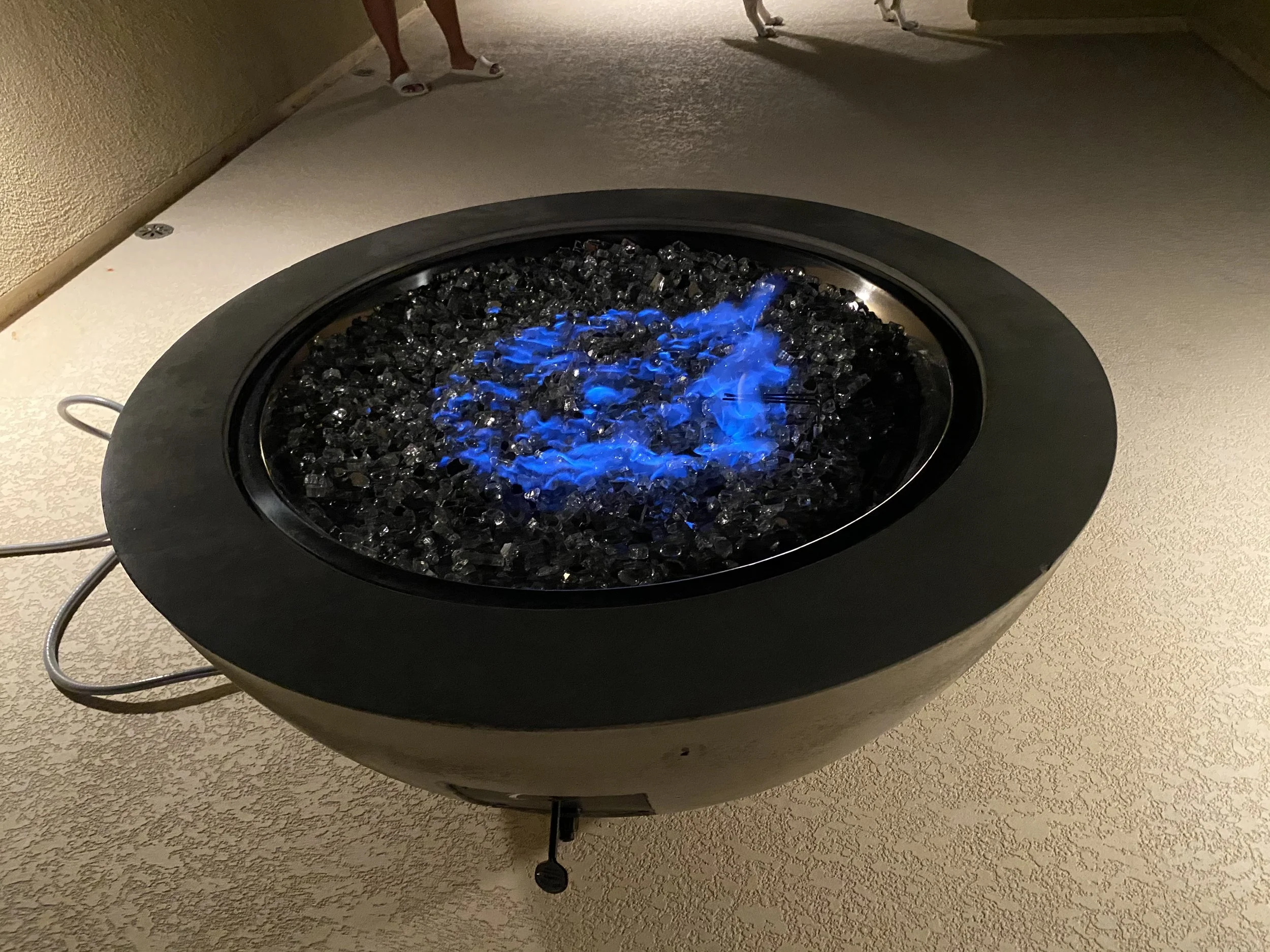 A round fire pit with blue flames and black glass stones on a textured surface.