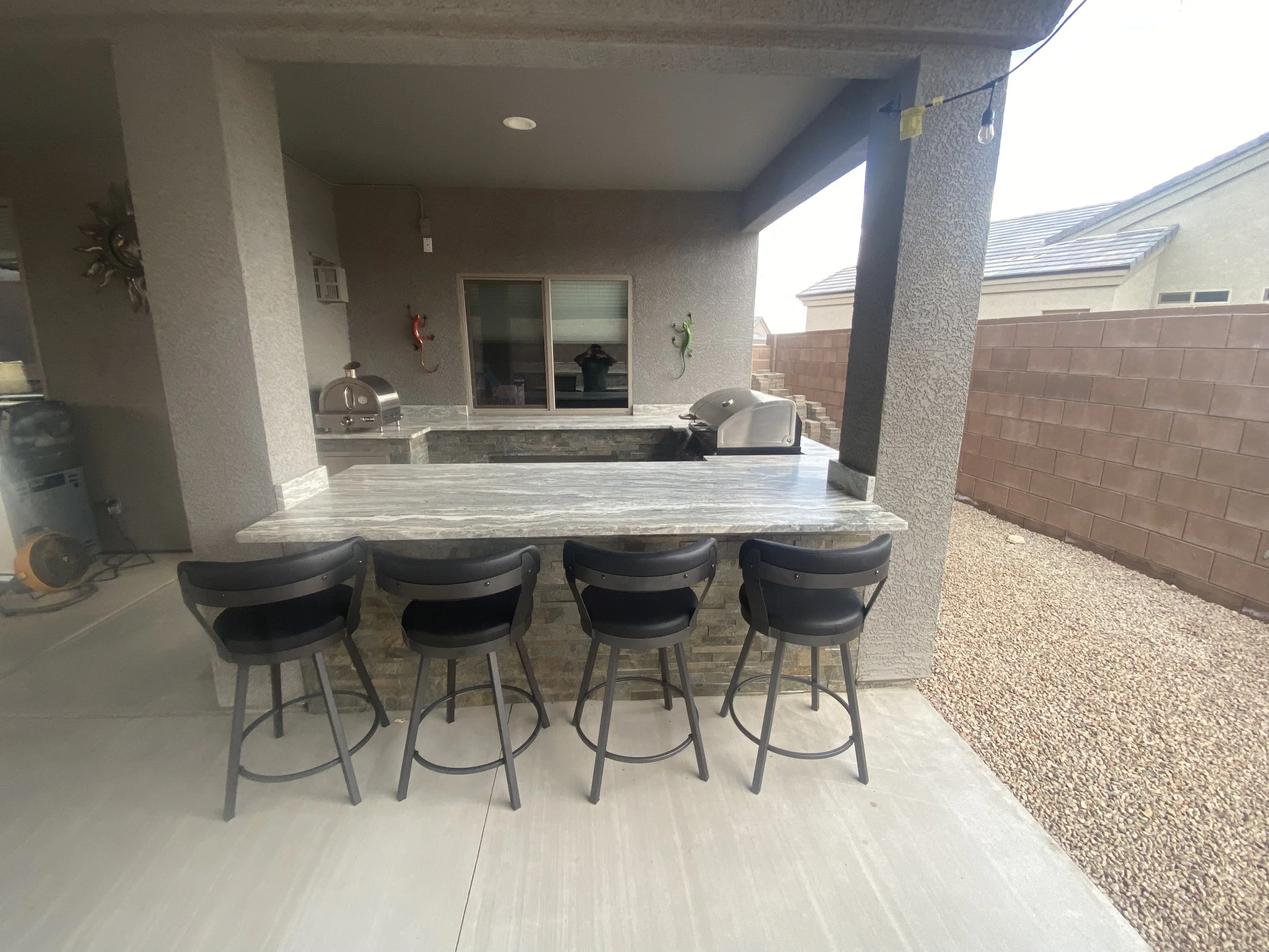 Outdoor kitchen with marble countertop, bar seating, grill, and pizza oven under a covered patio.
