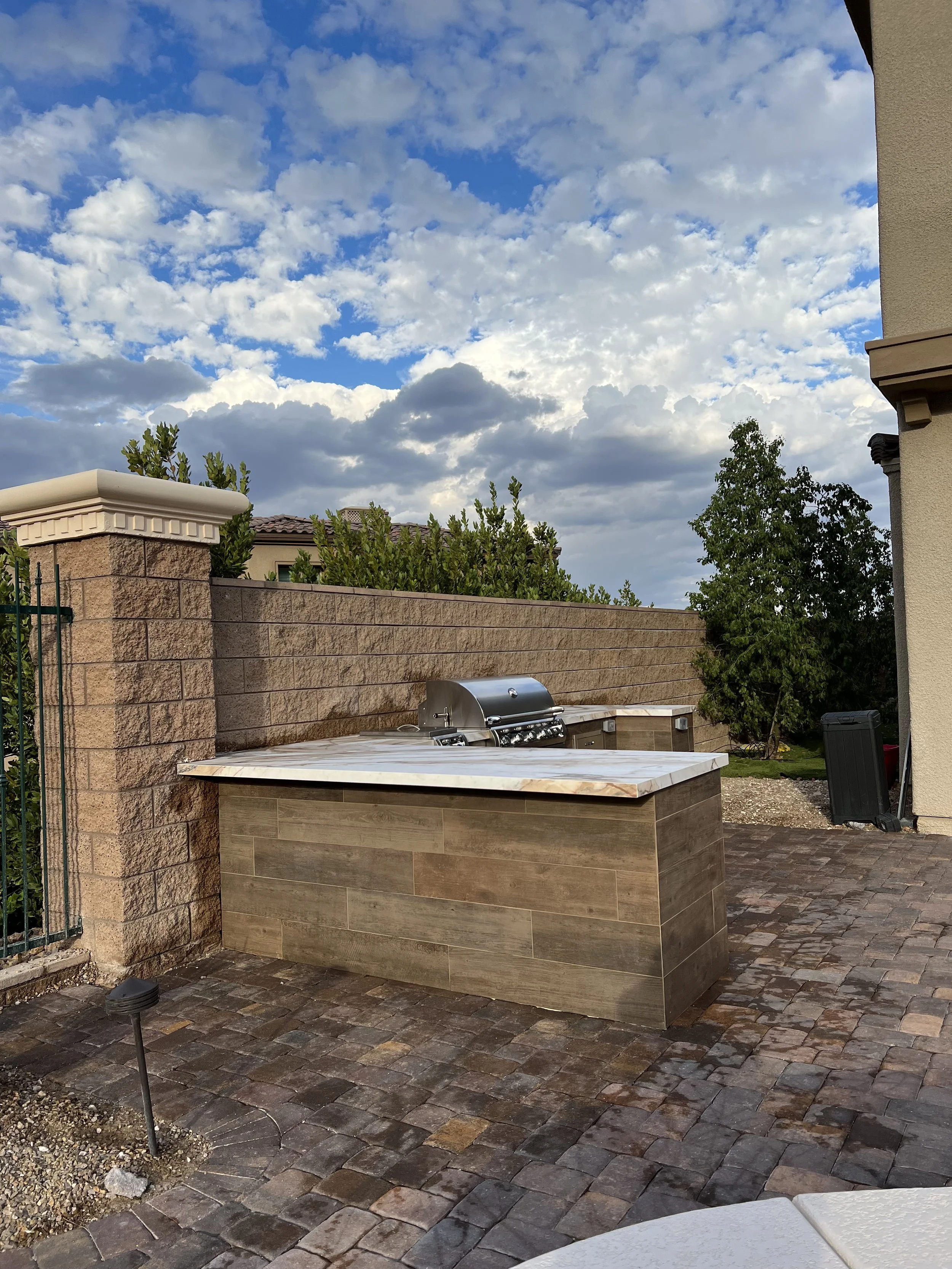 Outdoor kitchen area with built-in grill and stone countertop on a patio, surrounded by a stone wall and greenery, with a cloudy sky overhead.