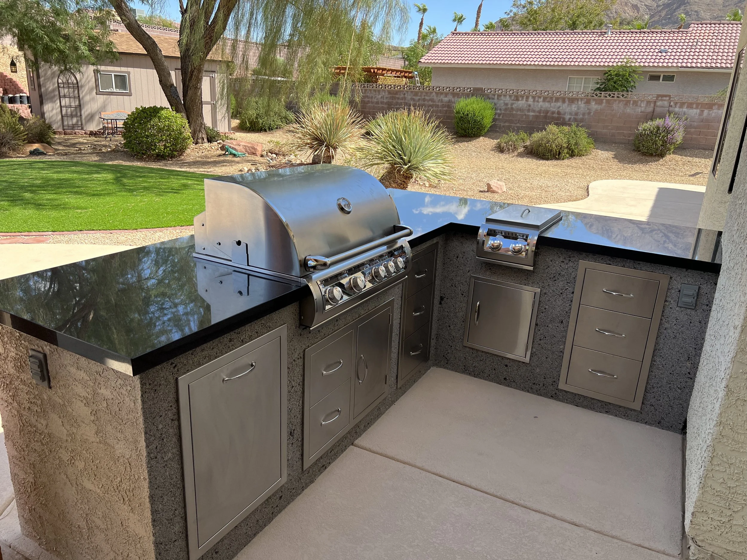 Outdoor kitchen with stainless steel grill and cabinets set in a backyard with landscaping, including trees and shrubs, against a background of houses.