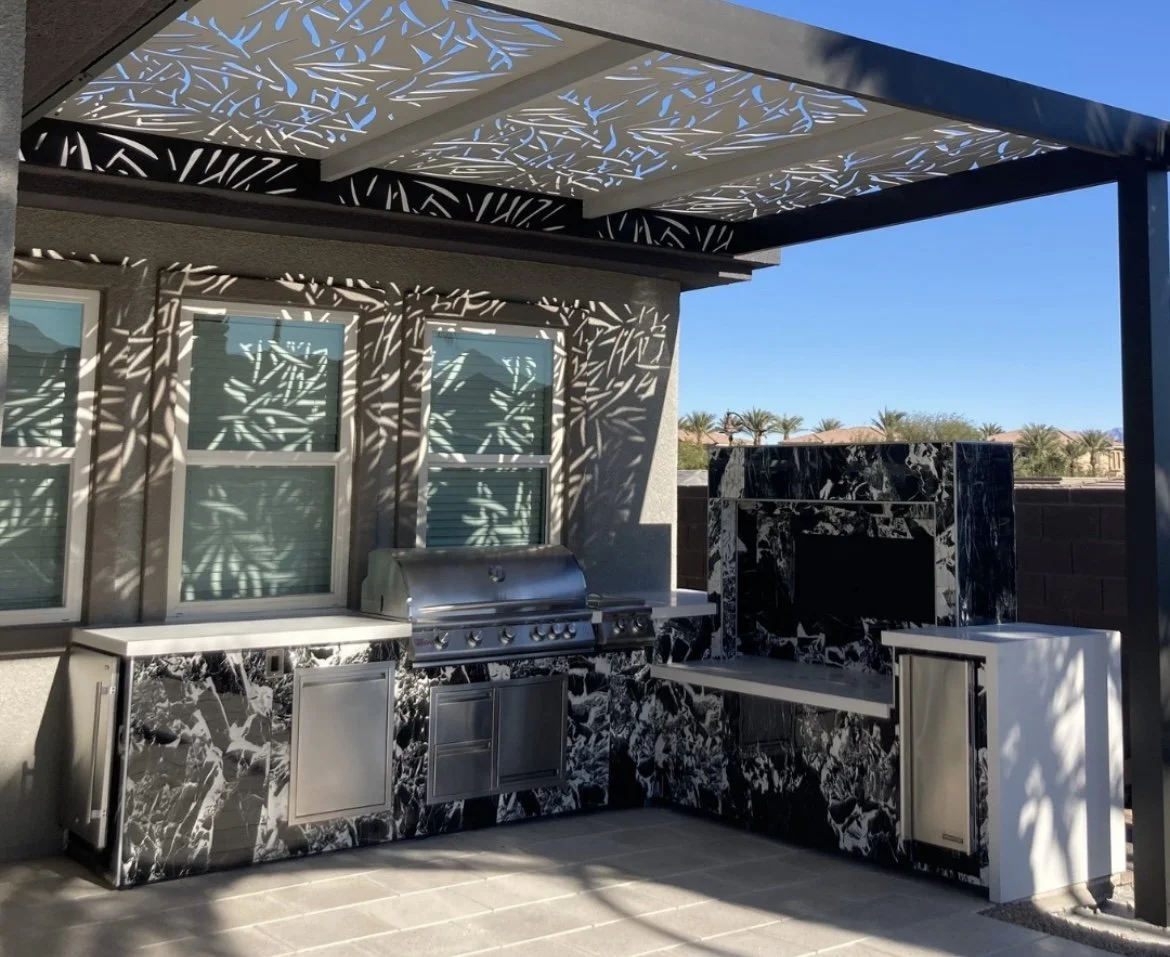 Outdoor kitchen with a stainless steel grill and marble countertops under a decorative pergola casting shadows.
