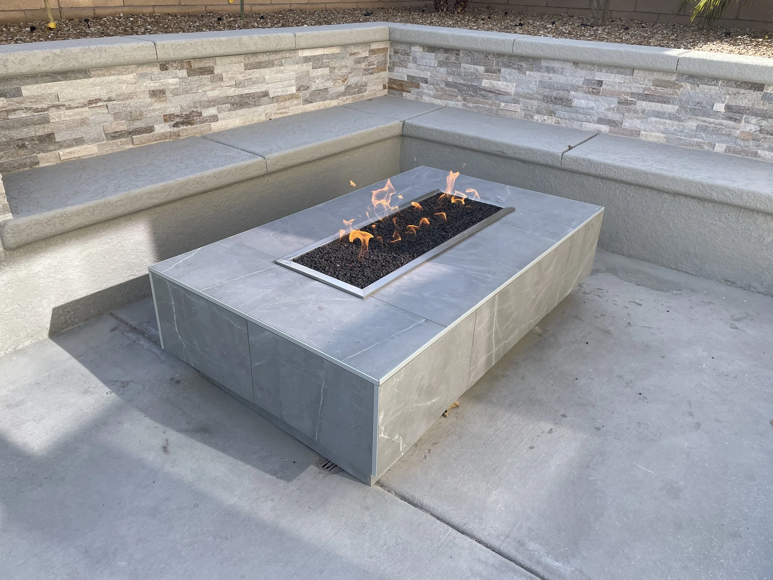 Modern outdoor fire pit with flames, tiled surface, stone bench, and stone wall backdrop.