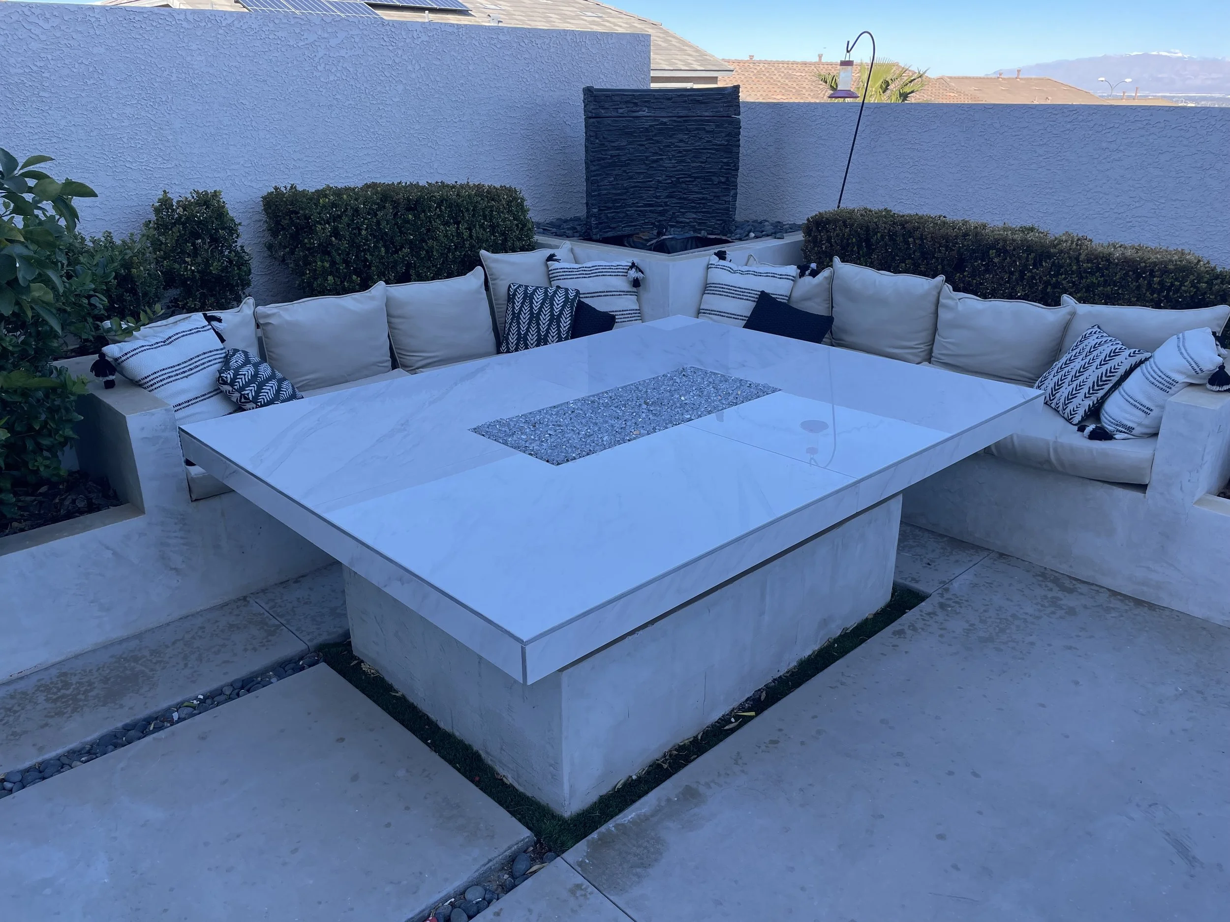 Outdoor patio with a white marble fire pit table, surrounded by L-shaped beige cushioned seating and decorative pillows. The area is enclosed by concrete walls and hedges.
