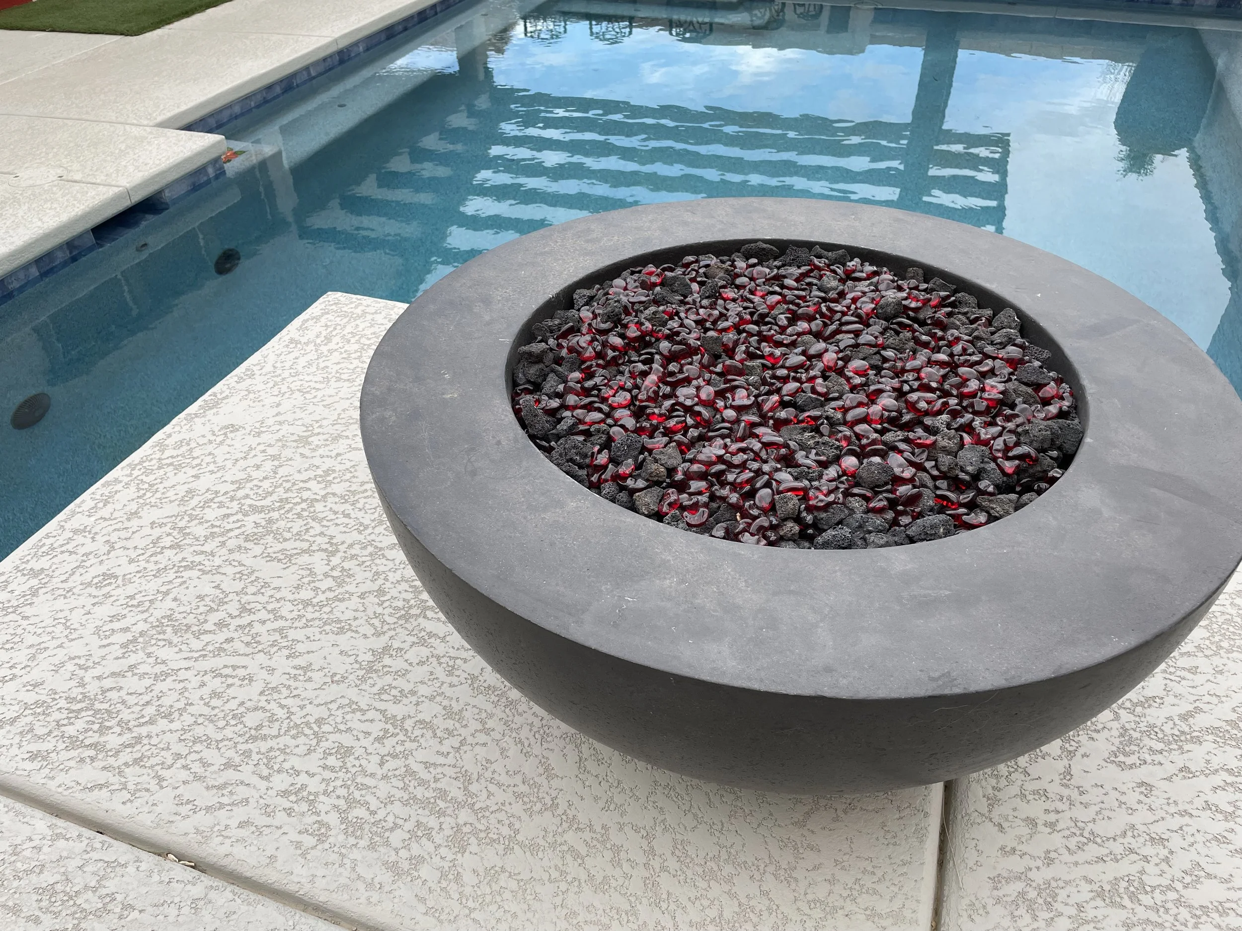 Outdoor fire pit with red stones and black lava rocks beside a swimming pool.