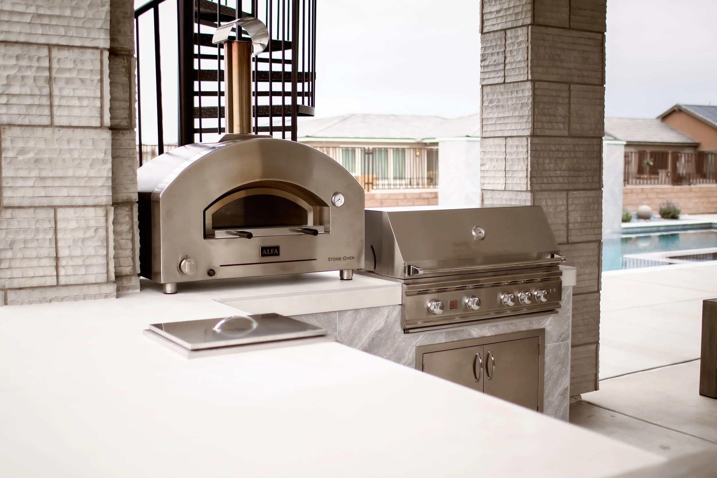 Outdoor kitchen with stainless steel pizza oven and grill next to a pool.