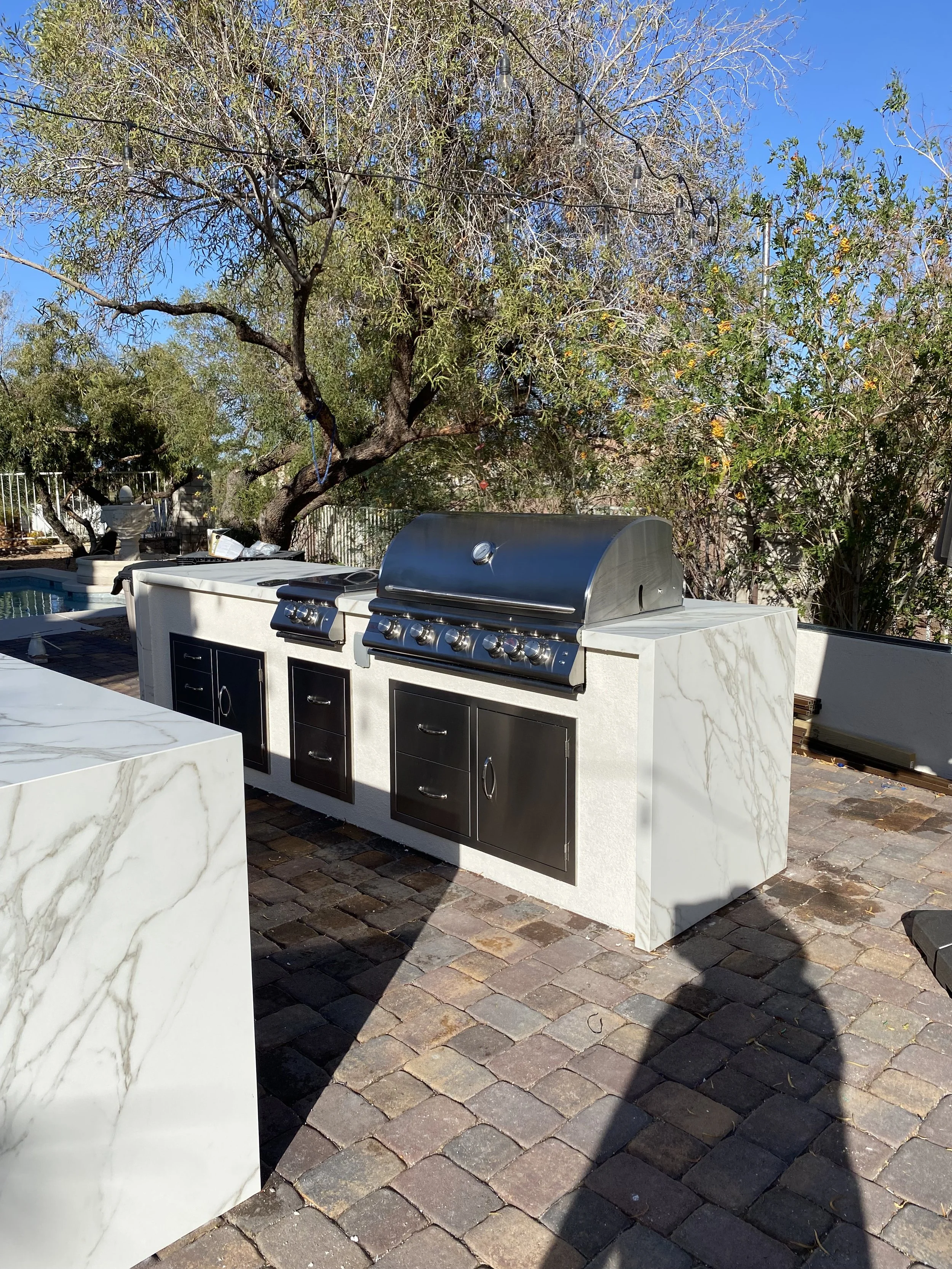 Outdoor grill station with stainless steel grills and marble countertops on a stone patio, surrounded by trees.