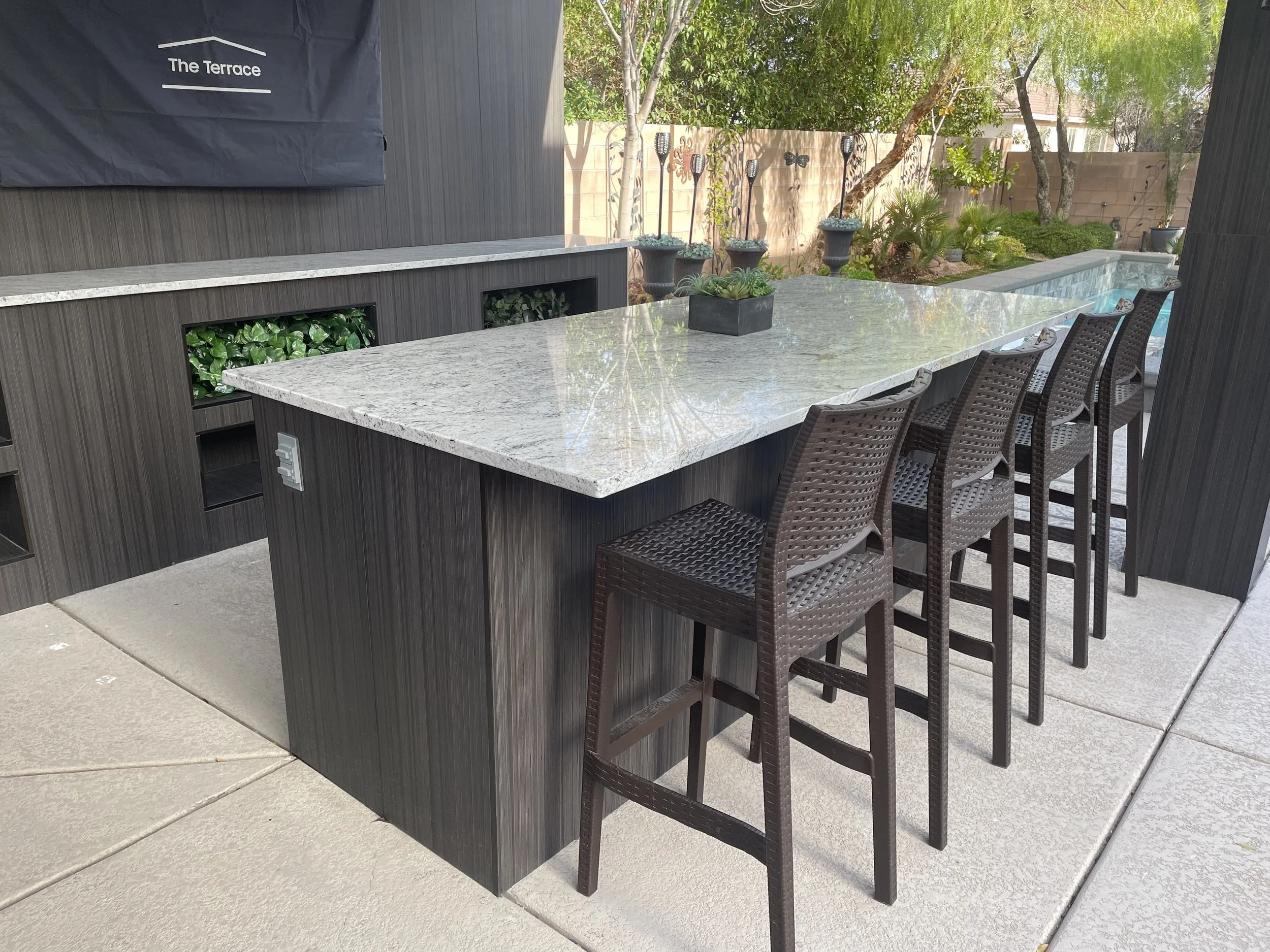 Outdoor patio bar with a marble countertop and wicker stools, surrounded by greenery and a small garden in the background.