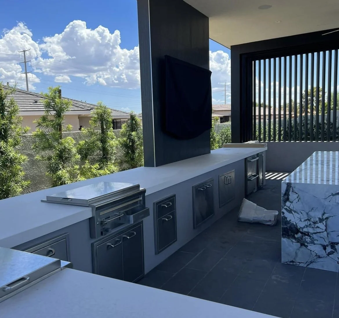 Outdoor kitchen with stainless steel appliances, a covered area, marble countertop, and a view of the sky with clouds and houses.