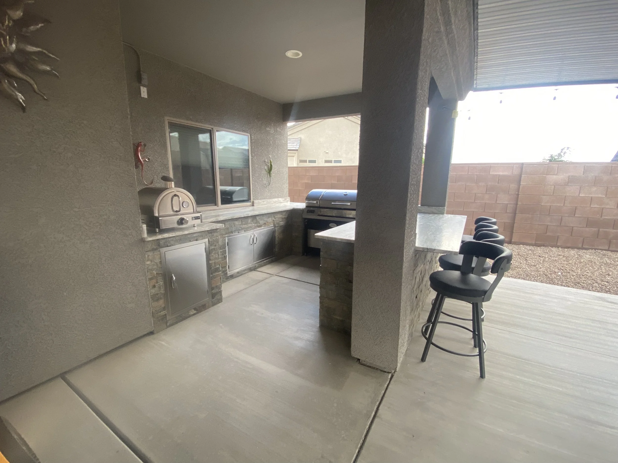 Outdoor kitchen with pizza oven, grill, countertop, barstools, and brick wall in backyard patio setting.