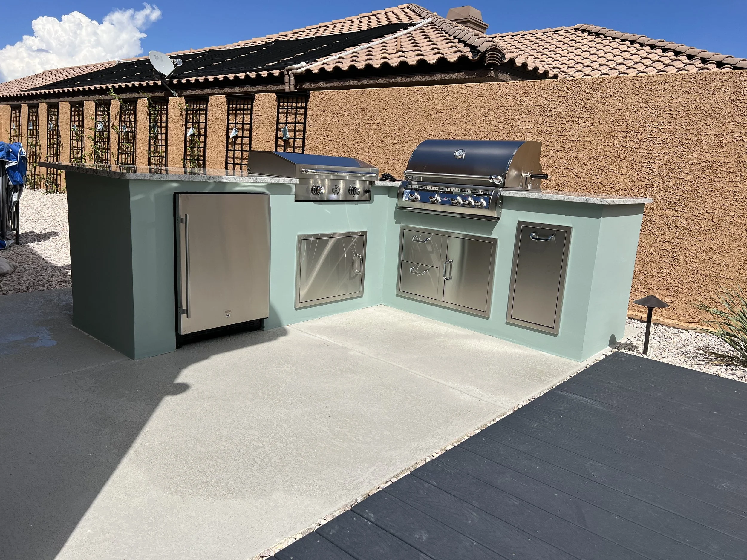 Outdoor kitchen with stainless steel grill, refrigerator, storage cabinets, and light blue cabinets, set against a brown stucco wall with a tiled roof in the background.