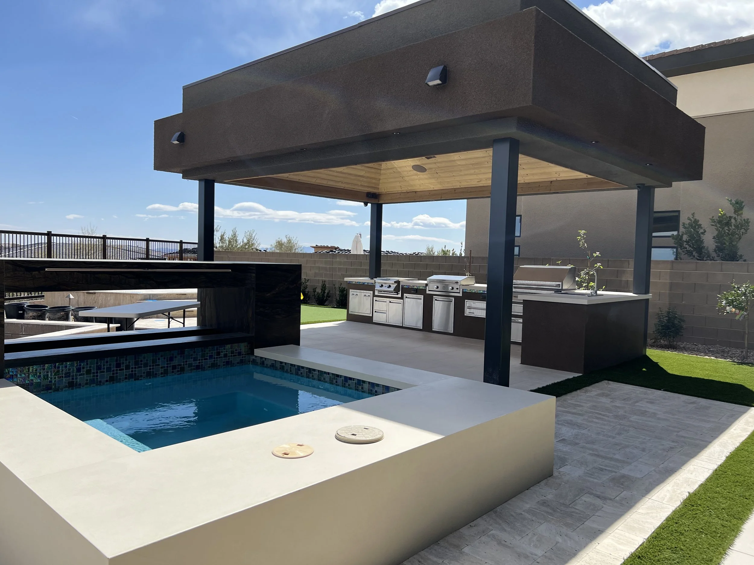 Outdoor kitchen with a covered patio, barbecue grills, and a small pool in a backyard setting.