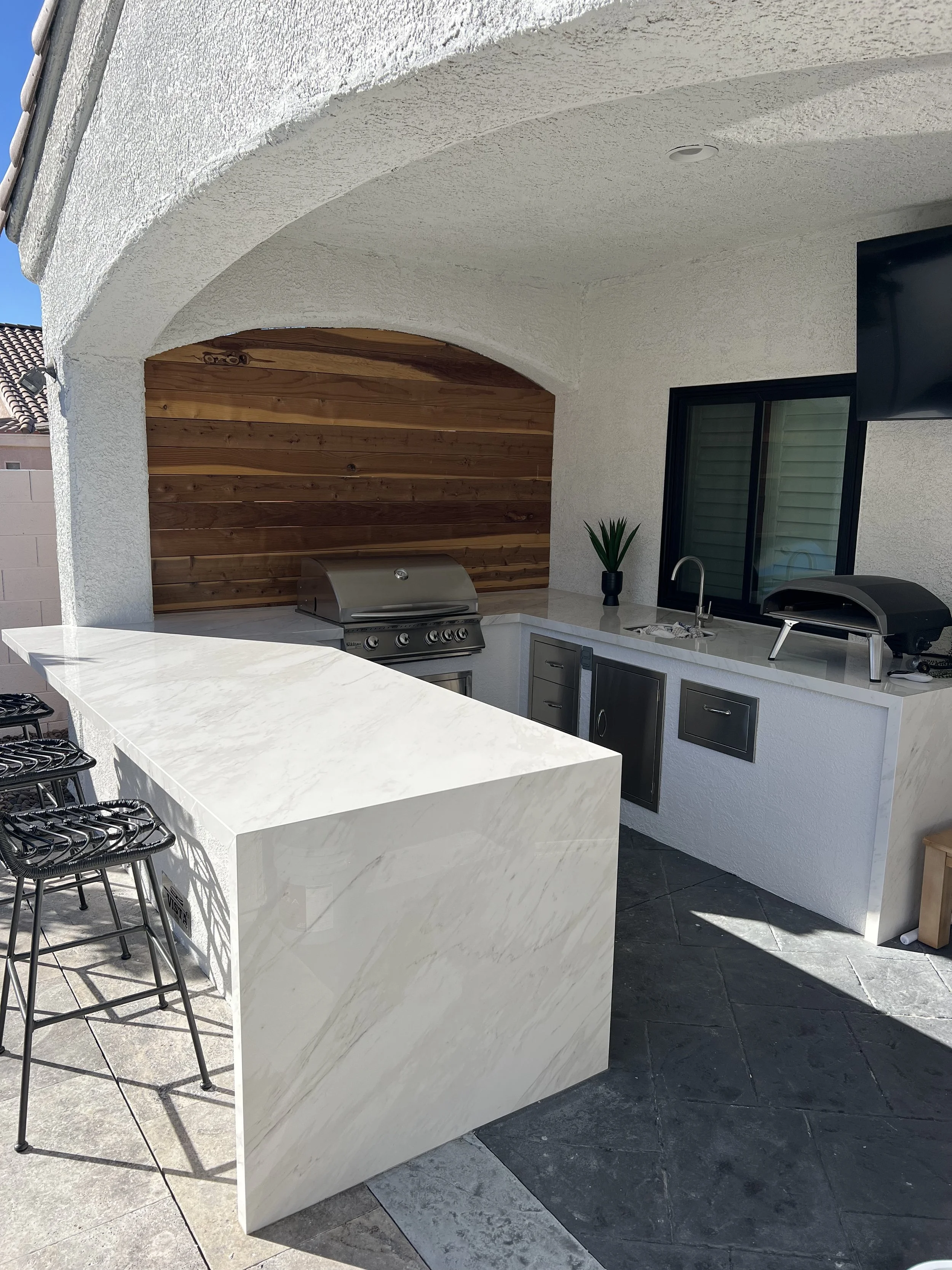 Outdoor kitchen with marble countertops, grill, and bar seating under a white textured arch.