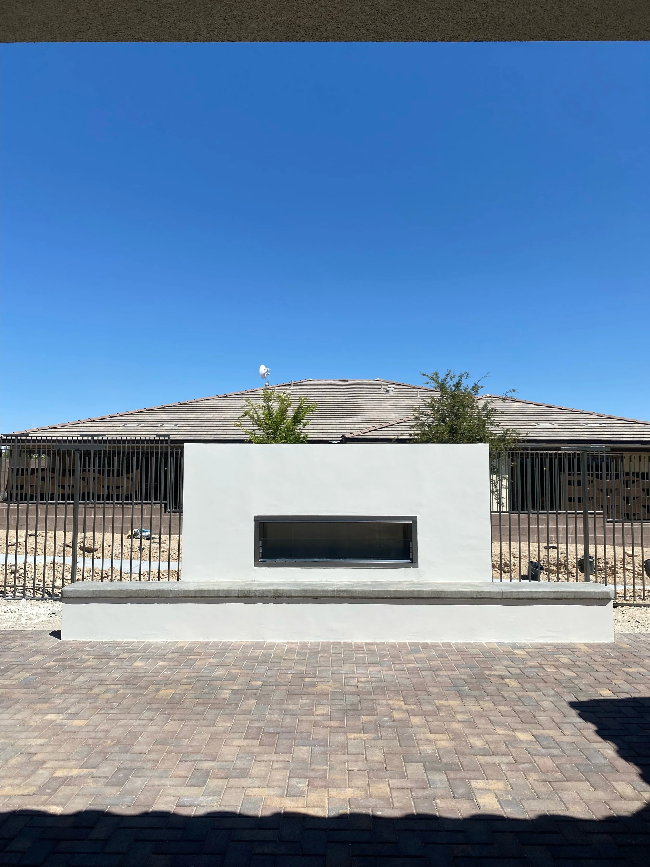 Outdoor fireplace in a backyard with a brick patio and metal fence, under a clear blue sky.