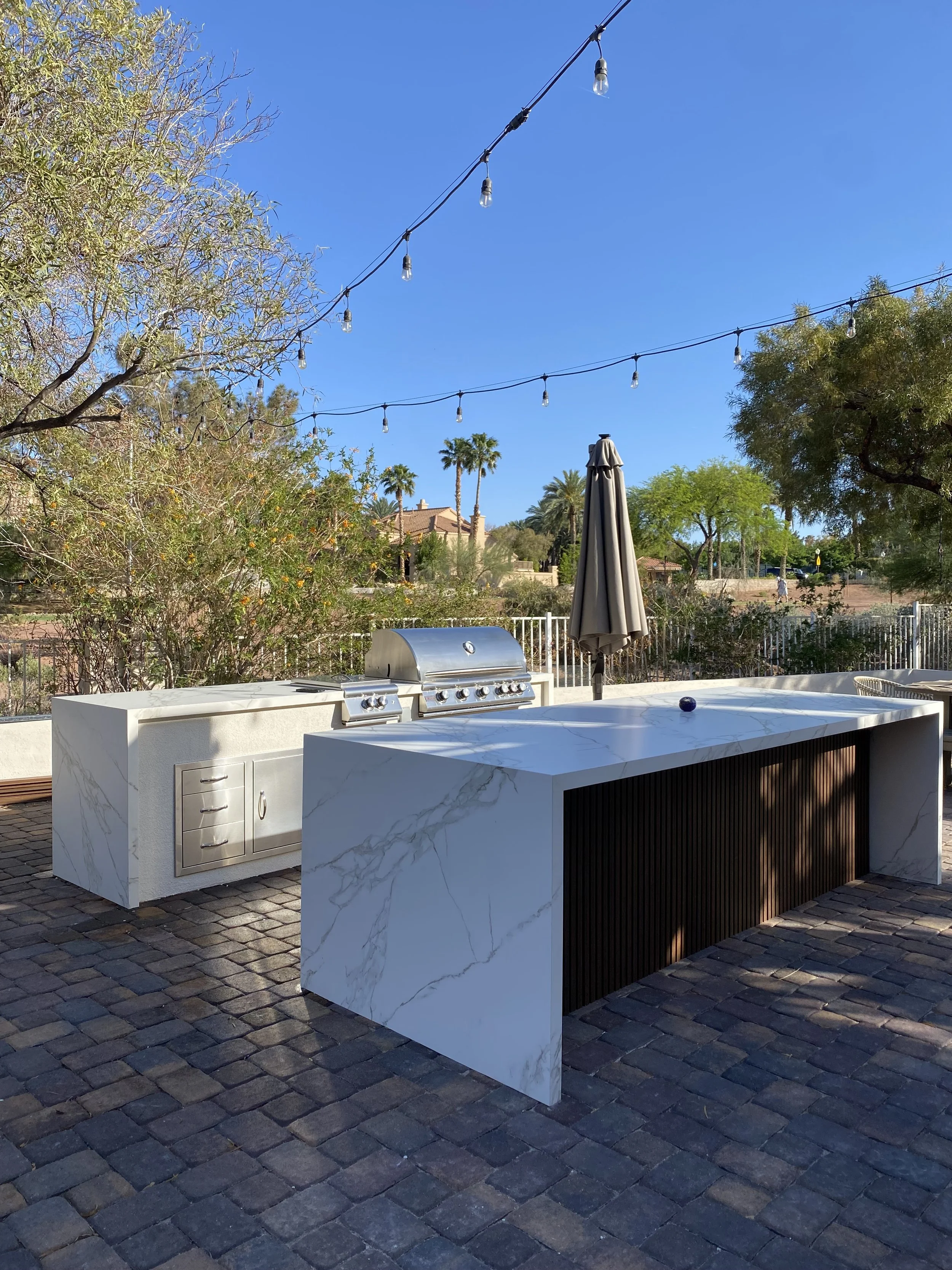 Outdoor kitchen area with a marble countertop and built-in stainless steel grill on a stone patio, surrounded by trees and string lights overhead.