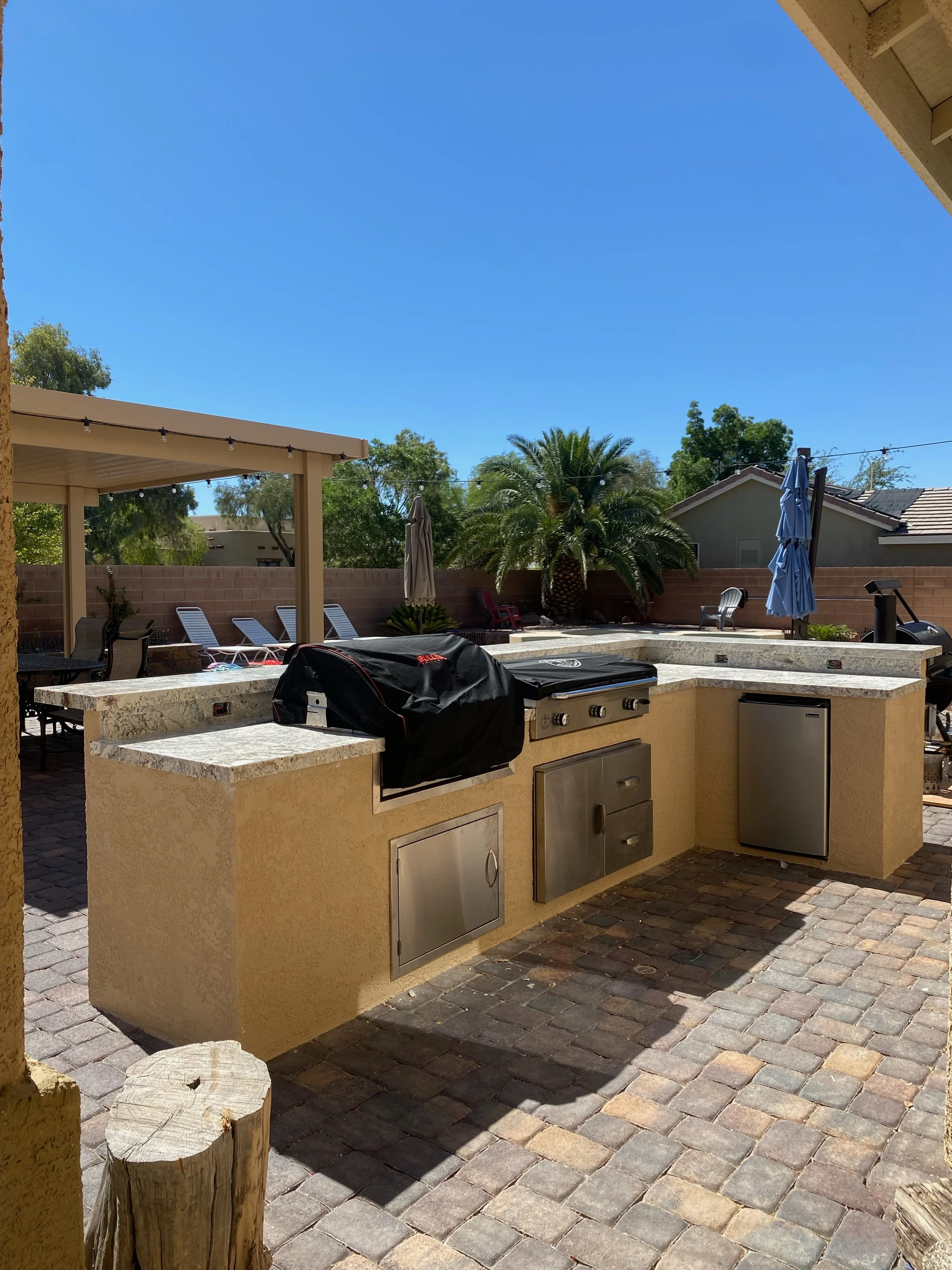 Outdoor patio with a built-in grill station, refrigerator, and seating area, surrounded by brick paving and green plants under a clear blue sky.