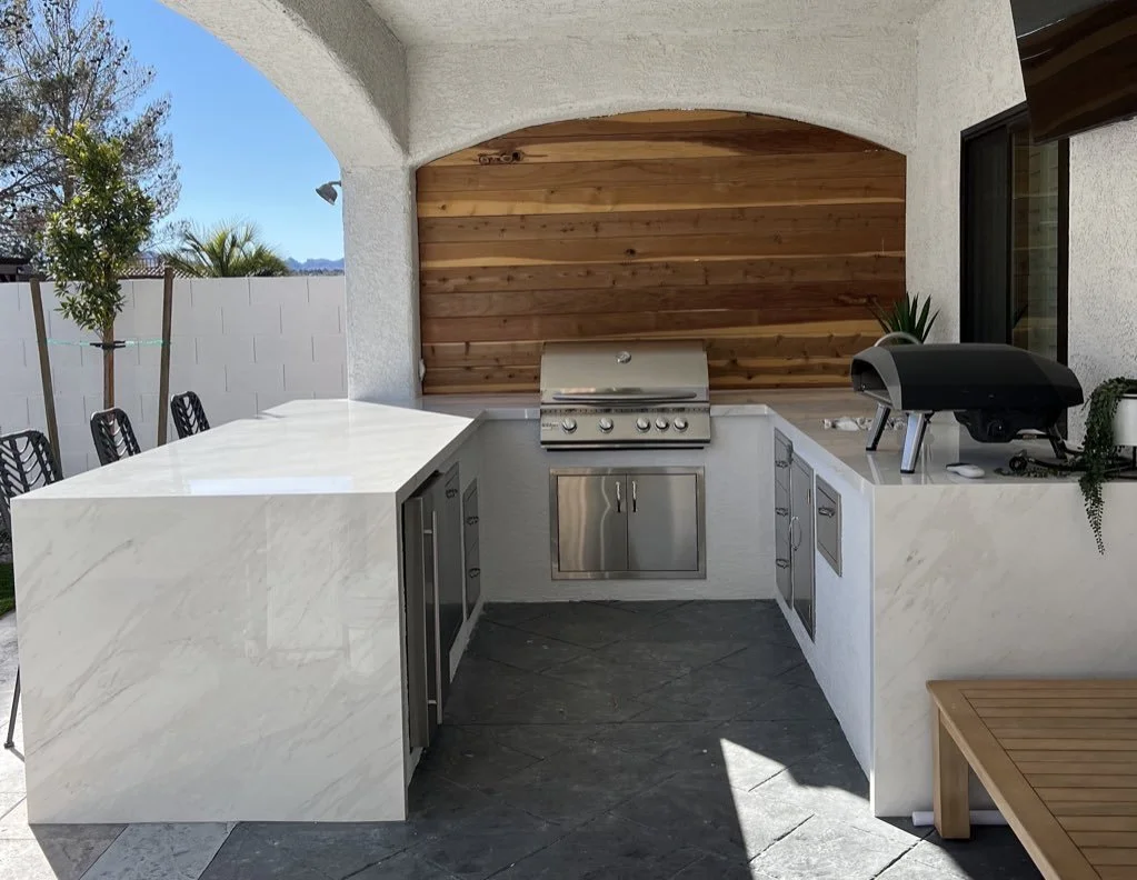 Outdoor kitchen with marble countertops, grill, and pizza oven under an arched covering.