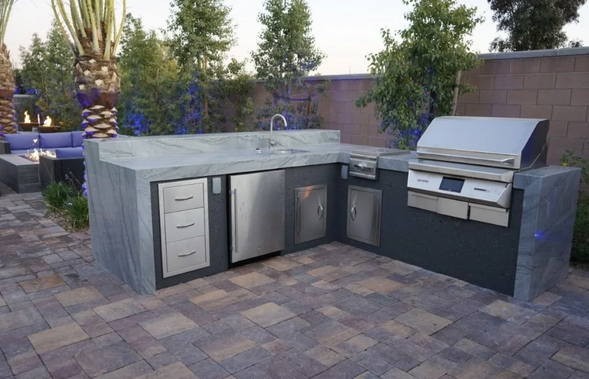 Outdoor kitchen with stainless steel appliances, including a grill and storage cabinets, on a stone patio surrounded by trees.
