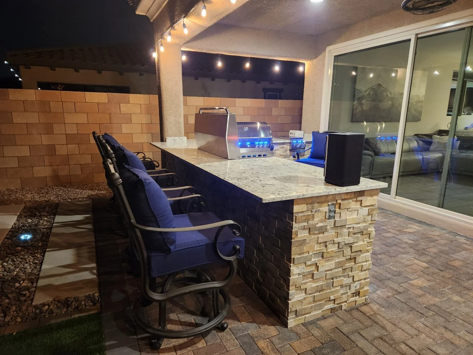 Outdoor kitchen with bar stools and a built-in grill, illuminated by string lights, on a patio with sliding glass doors and a brick wall.