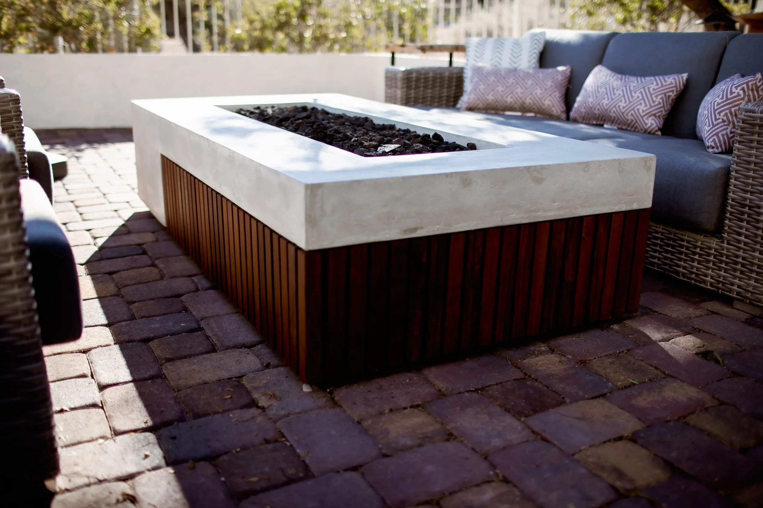Outdoor patio with a modern firepit on a brick surface, surrounded by wicker furniture and cushioned seating.