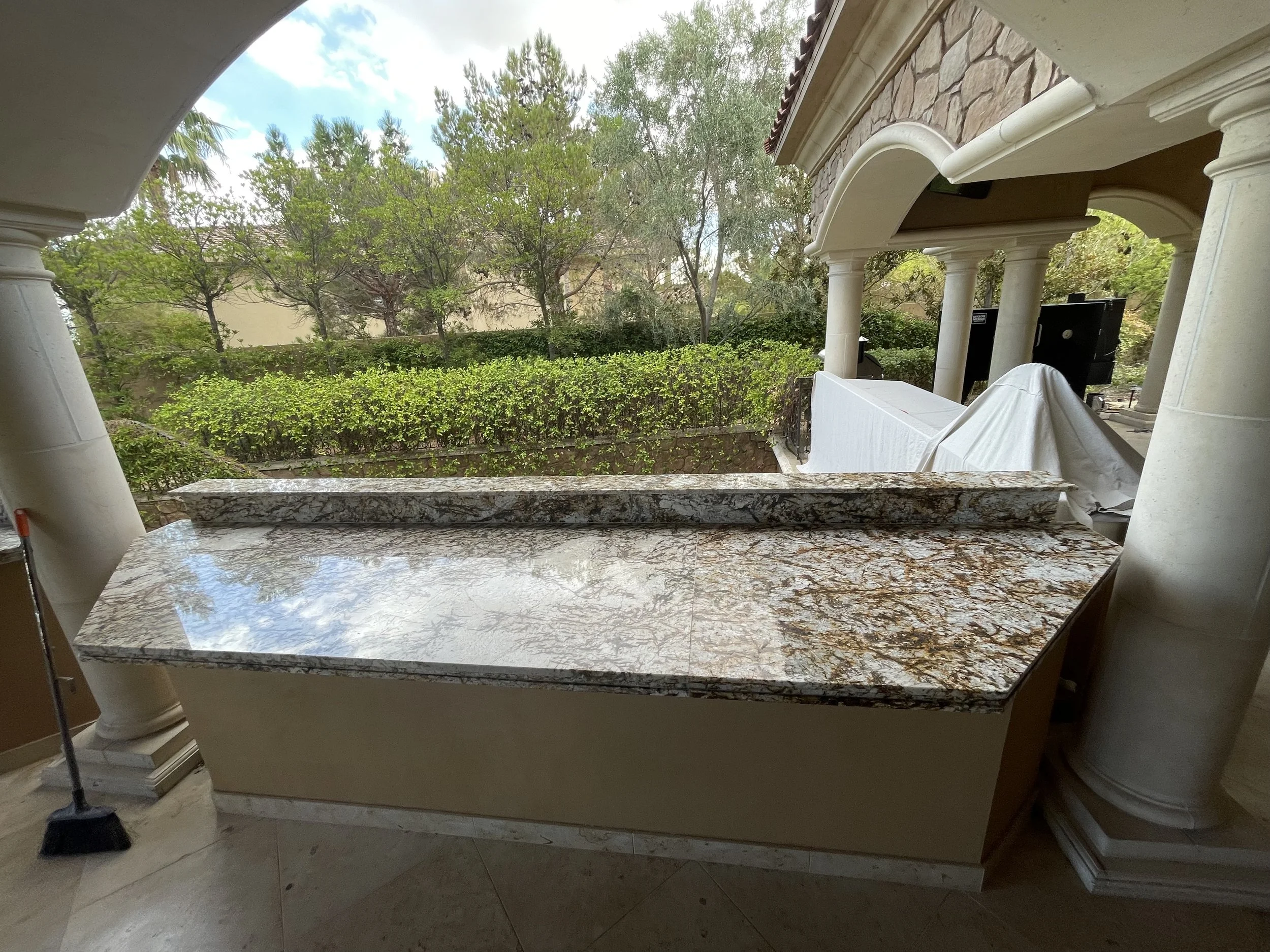 Outdoor patio with granite countertop and stone columns, view of trees and bushes in background.