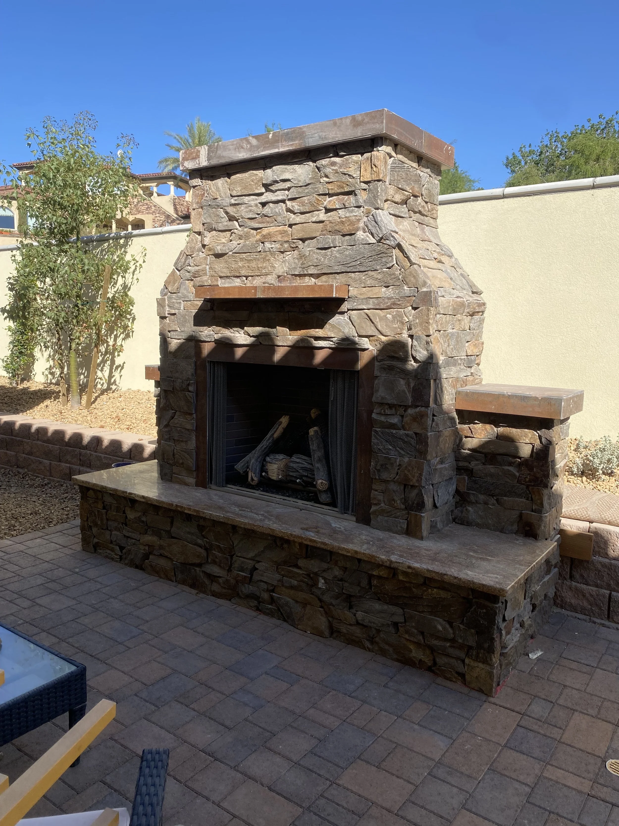 Outdoor stone fireplace on patio with brick flooring, surrounded by trees and a residential building in the background.