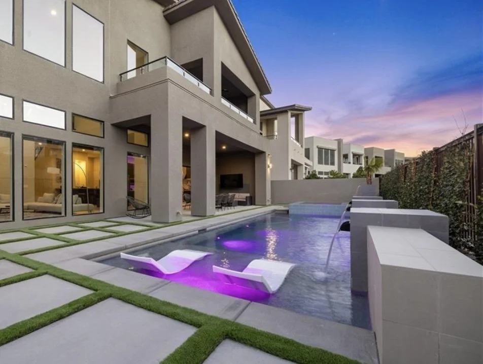 Modern house exterior with large windows and a spacious patio featuring a sleek swimming pool with water features and lounge chairs, set against a twilight sky.